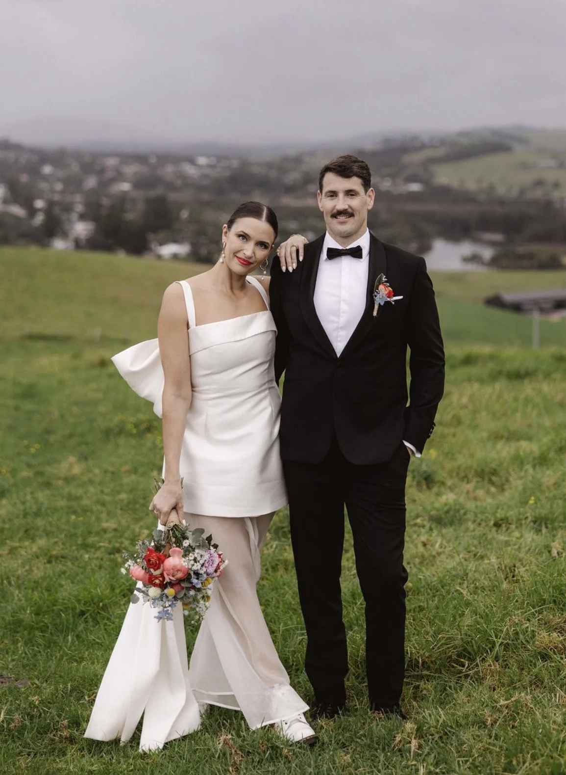 A bride and groom standing outdoors on a grassy field, celebrating their wedding. The bride wears a white dress with a bow on her back, holding a colorful bouquet, and the groom wears a black tuxedo with a bow tie, smiling and resting her hand on his shoulder.