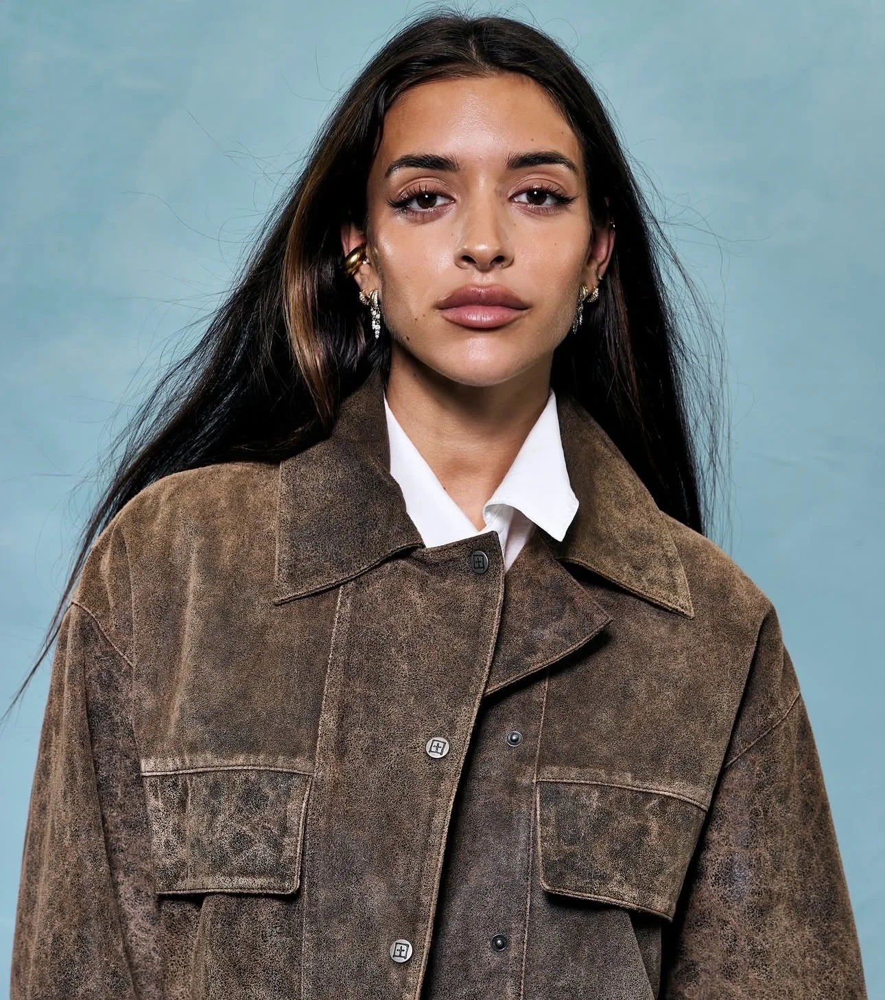 A woman with long dark hair, wearing earrings, a white shirt, and a brown jacket, posing against a light blue background.