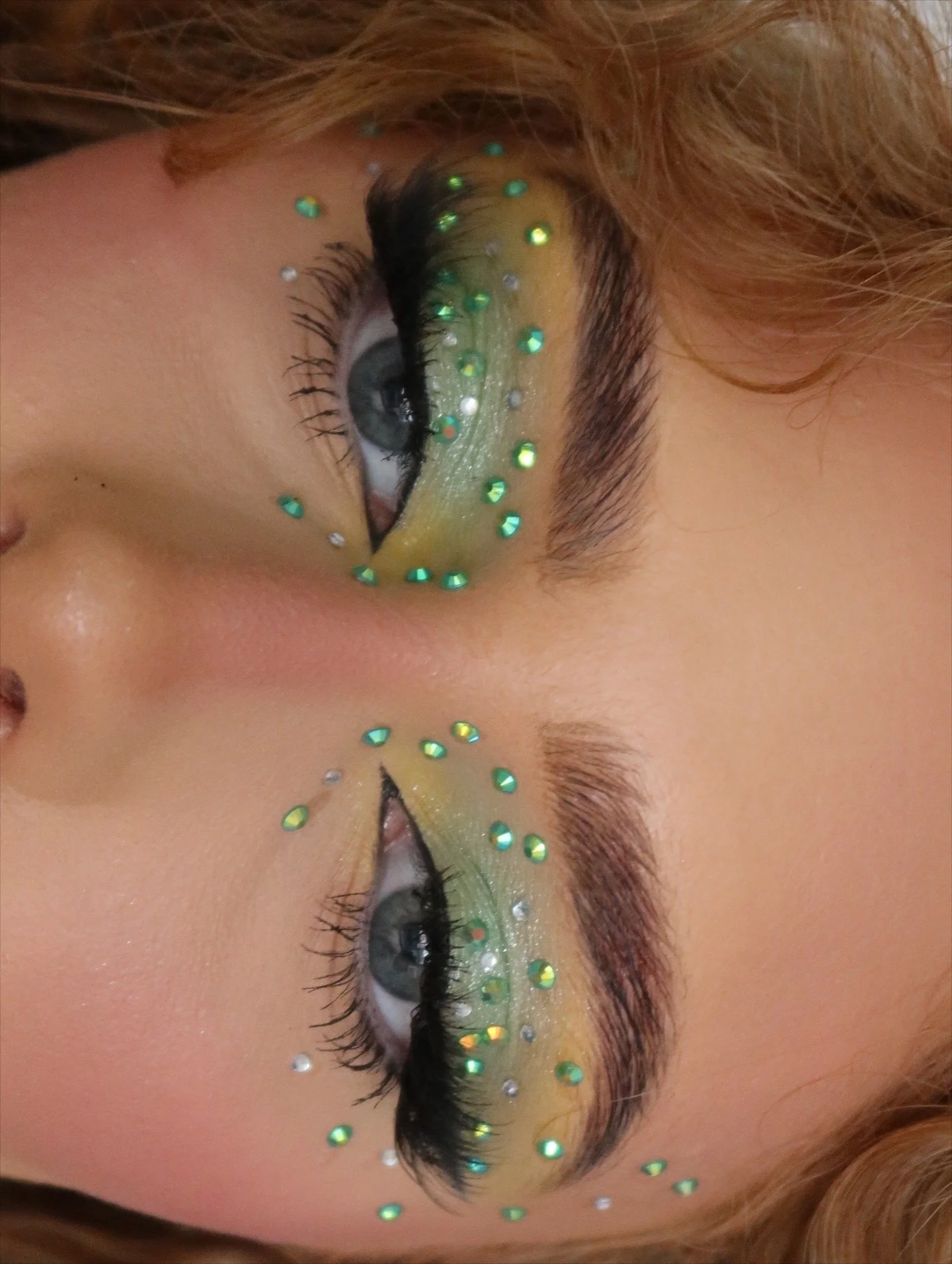 Close-up of a person's eye with eye makeup featuring yellow eyeshadow and decorative rhinestones, with the image flipped upside down.