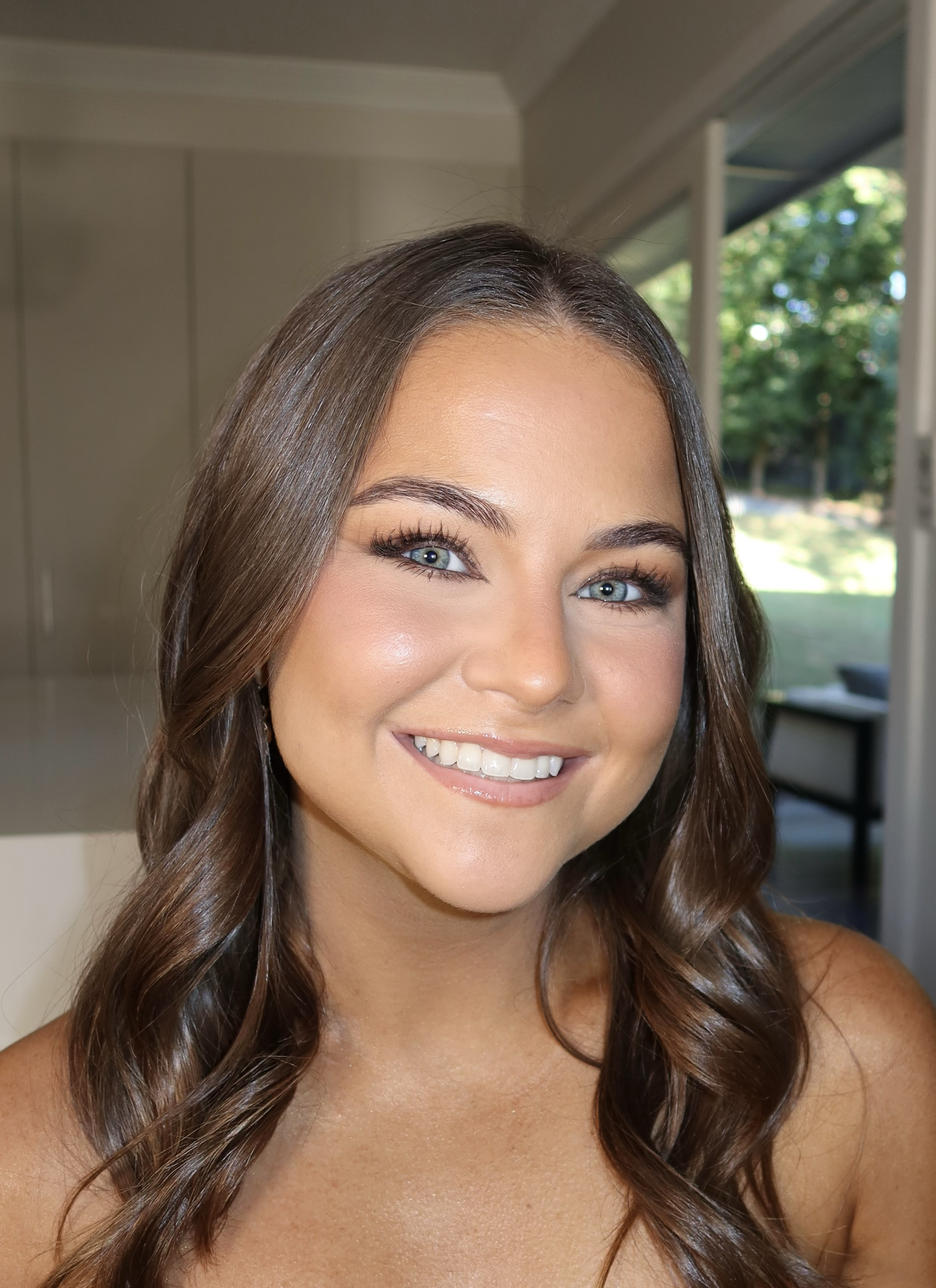 A smiling woman with long wavy brown hair, wearing makeup, and posing indoors near a sliding glass door that shows greenery outside.