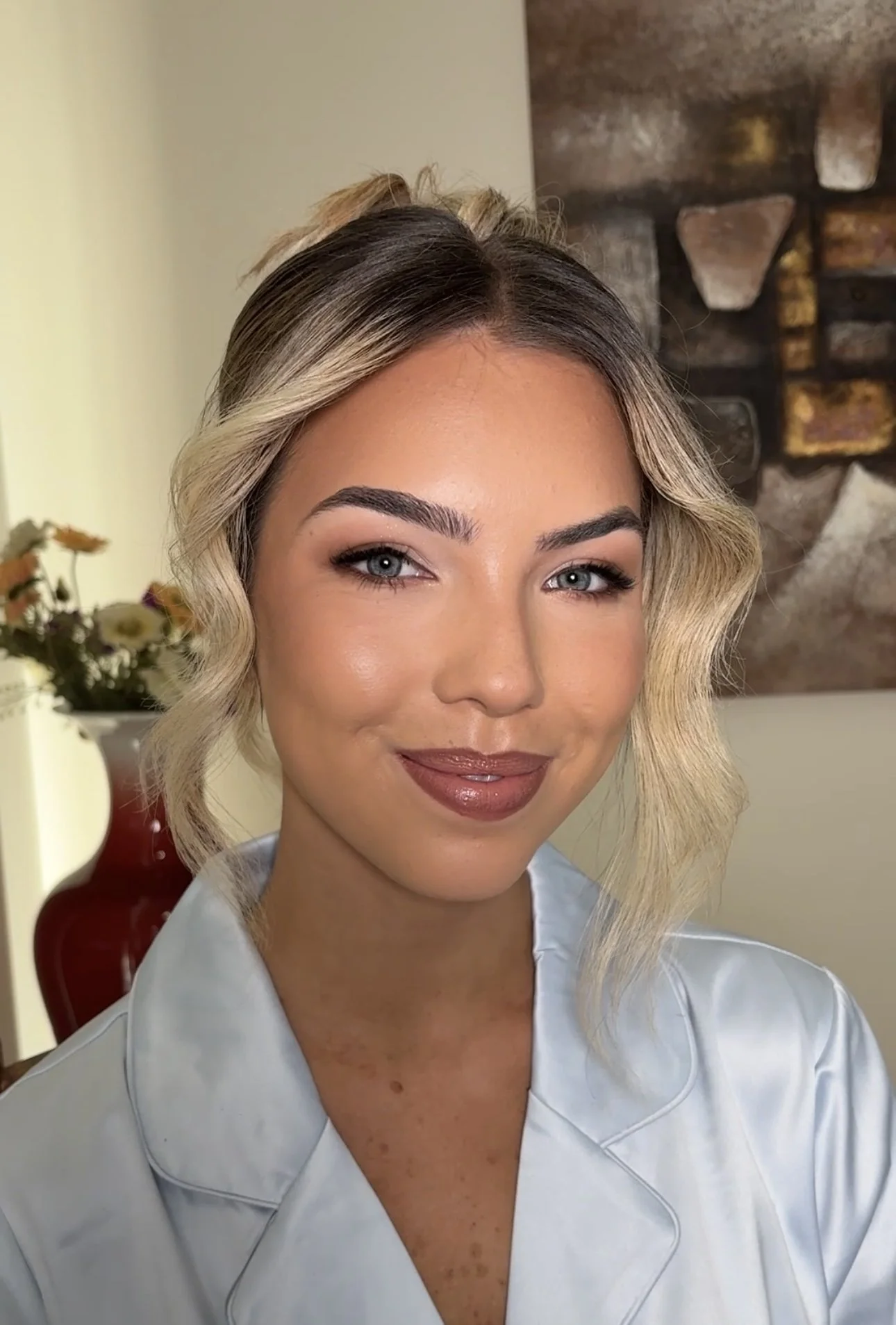 A woman with blonde hair styled in waves, wearing makeup, and a light-colored satin top, posing indoors with a decorative houseplant and a stone wall in the background.
