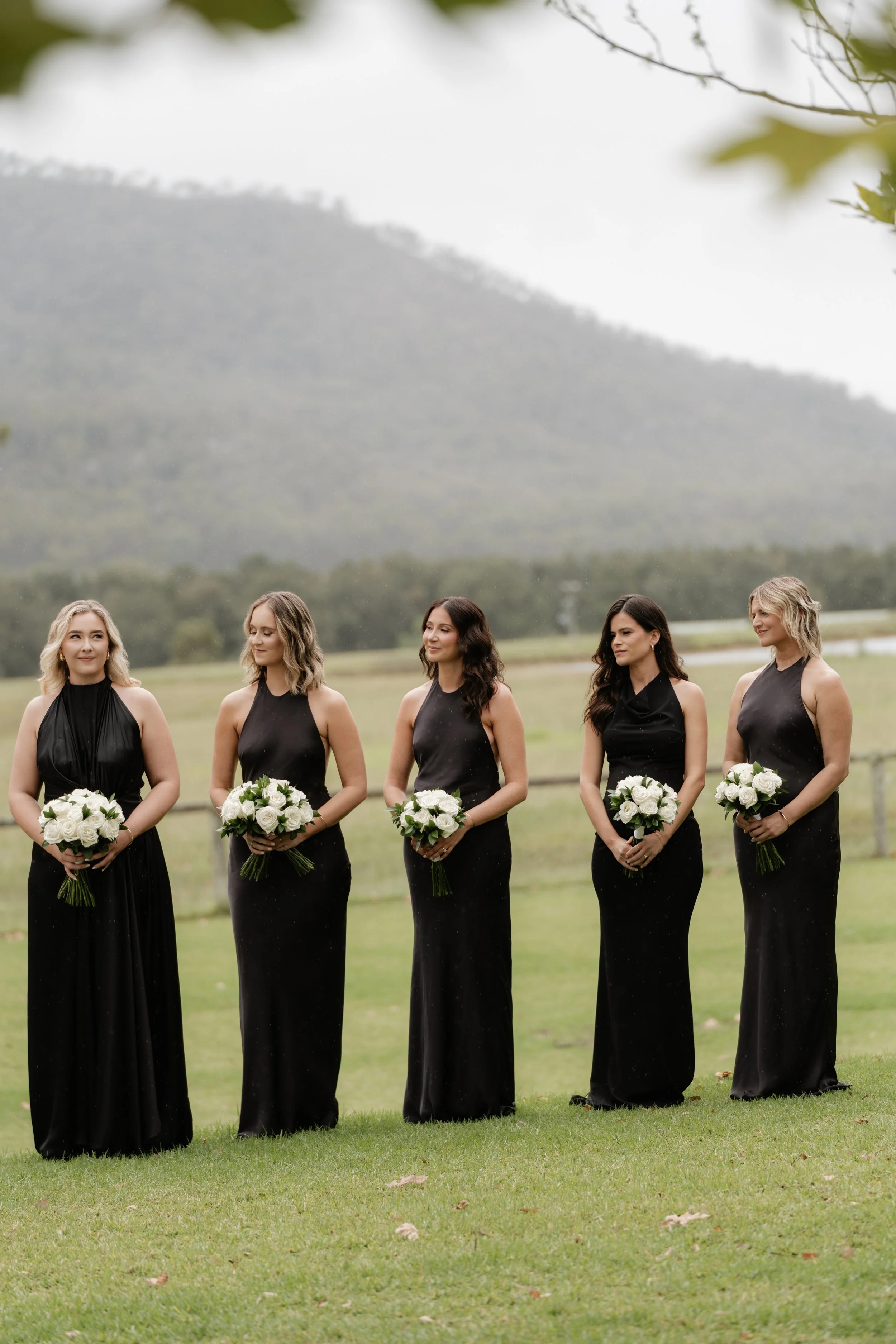 Five women dressed in black evening gowns, holding white flower bouquets, standing outdoors on a grassy field with a mountain and cloudy sky in the background.