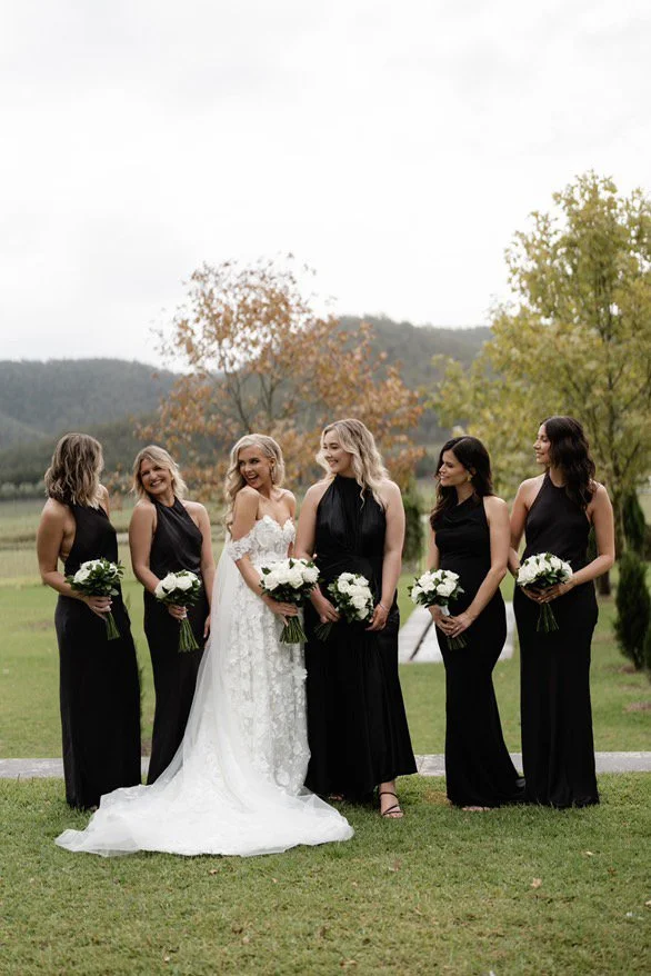 A bride in a white wedding dress standing with five bridesmaids in black dresses, all holding white bouquets outdoors with trees and mountains in the background.