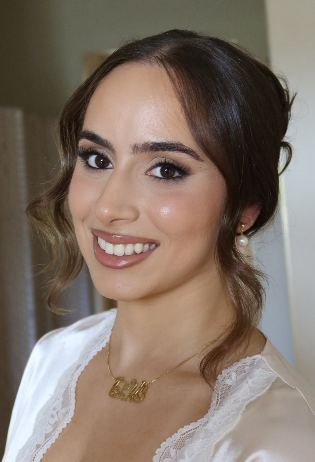 A young woman with brown hair styled in loose waves, wearing pearl earrings and a gold necklace that reads 'Tina'. She has makeup with defined eyebrows, eyeliner, blush, and glossy lipstick. She is smiling and wearing a white lace-trimmed top.