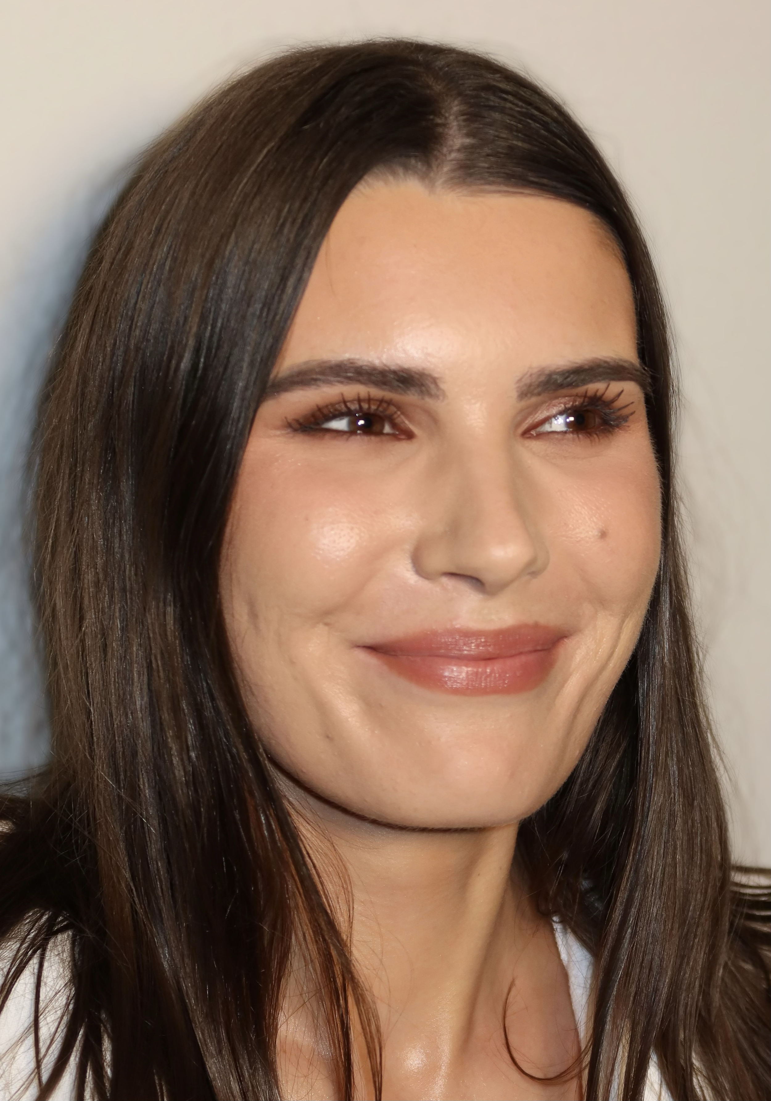 Close-up of a woman with long dark hair, smiling with bright eyes and wearing makeup.