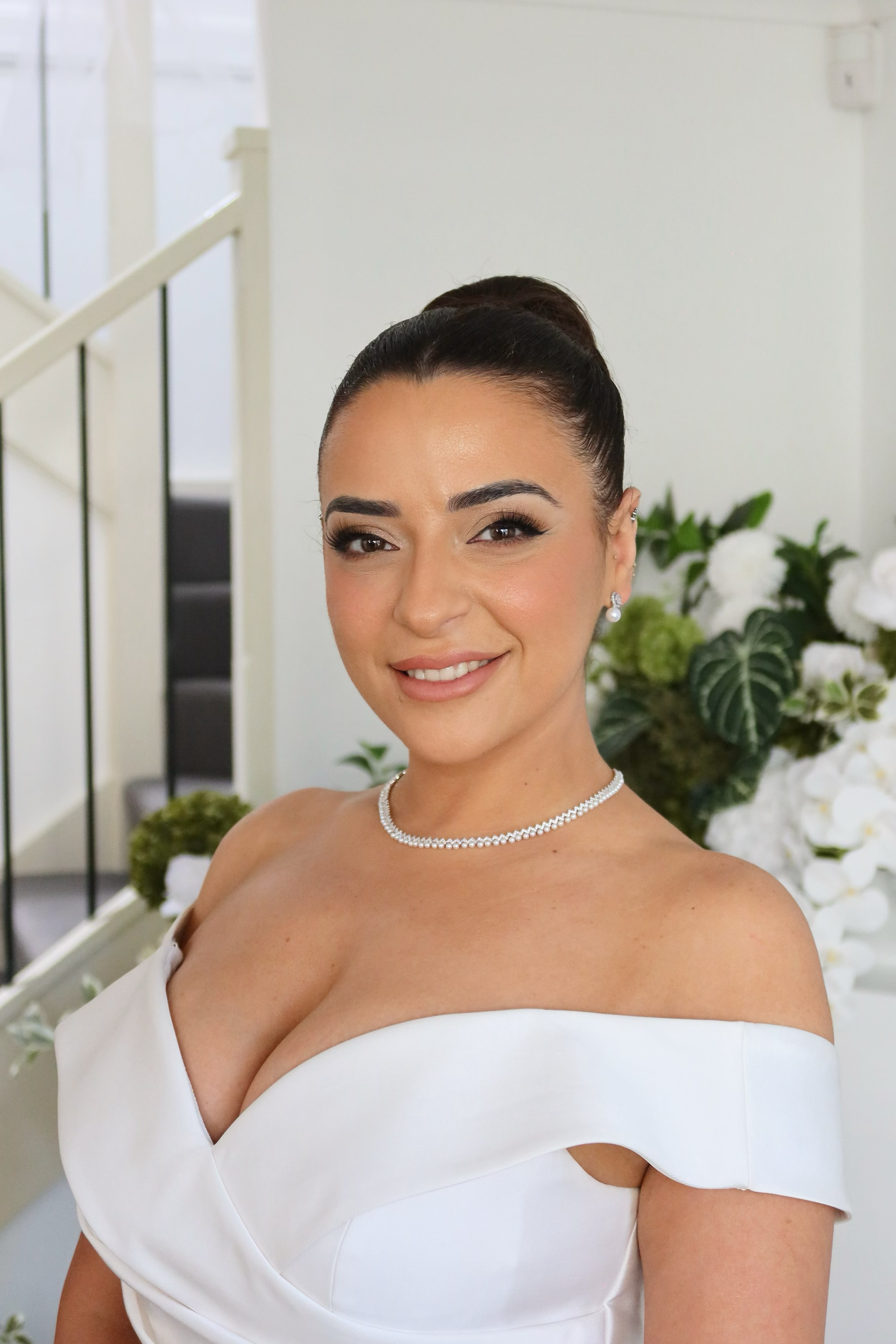 A woman in a white off-the-shoulder dress, wearing pearl jewelry, smiling and winking indoors with a staircase and floral arrangement in the background.