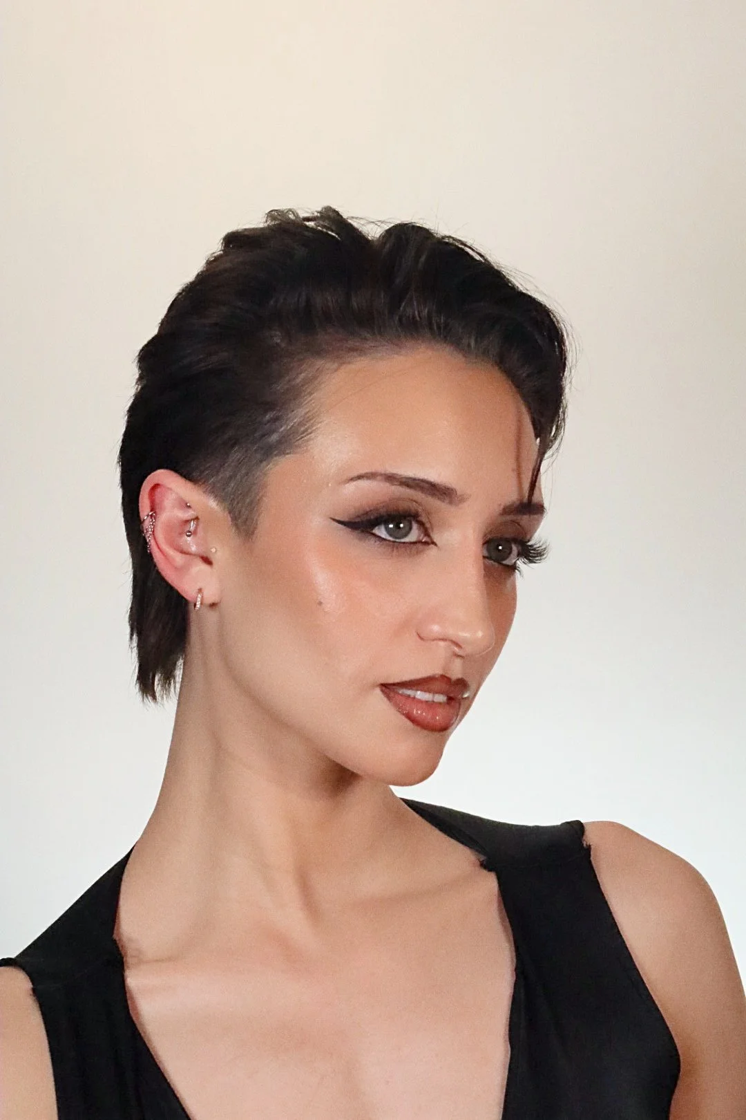A woman with short, dark, styled hair, wearing makeup with winged eyeliner, and earrings, dressed in a sleeveless black top, against a plain light background.