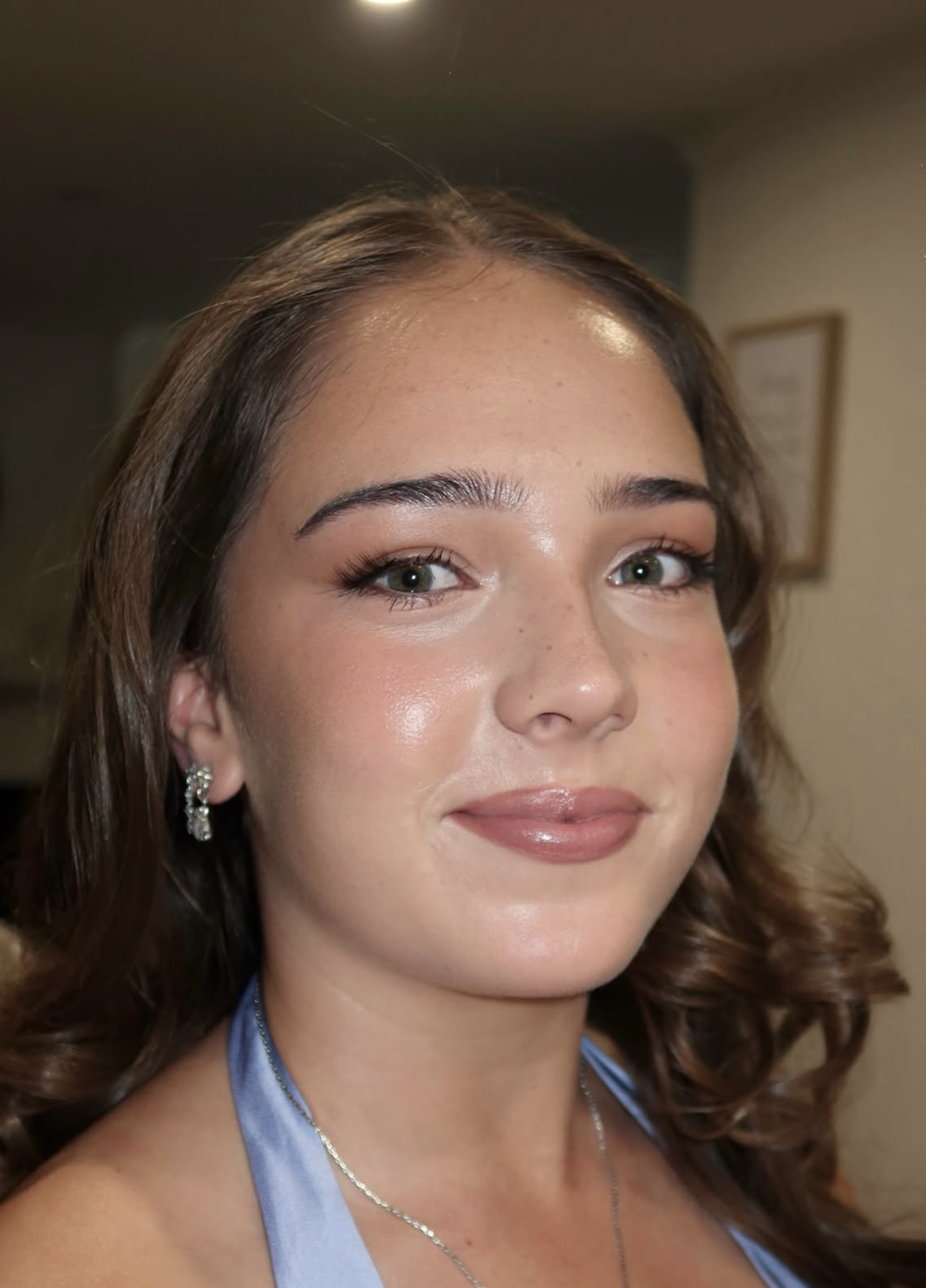 A young woman with brown, curly hair, blue eyes, and makeup, wearing earrings and a light blue top, smiling in an indoor setting.