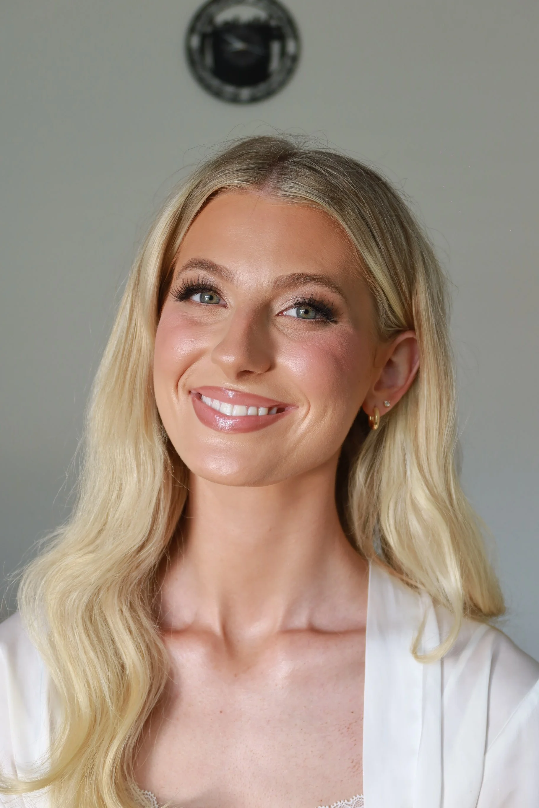 Close-up of a smiling woman with long blonde hair, wearing makeup and gold earrings, in front of a plain background with a security camera overhead.