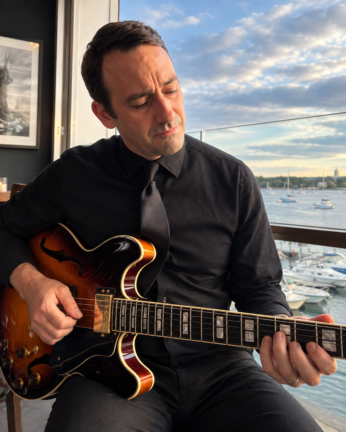 A man in a black shirt and tie playing an electric guitar near a window overlooking a marina with boats and water.