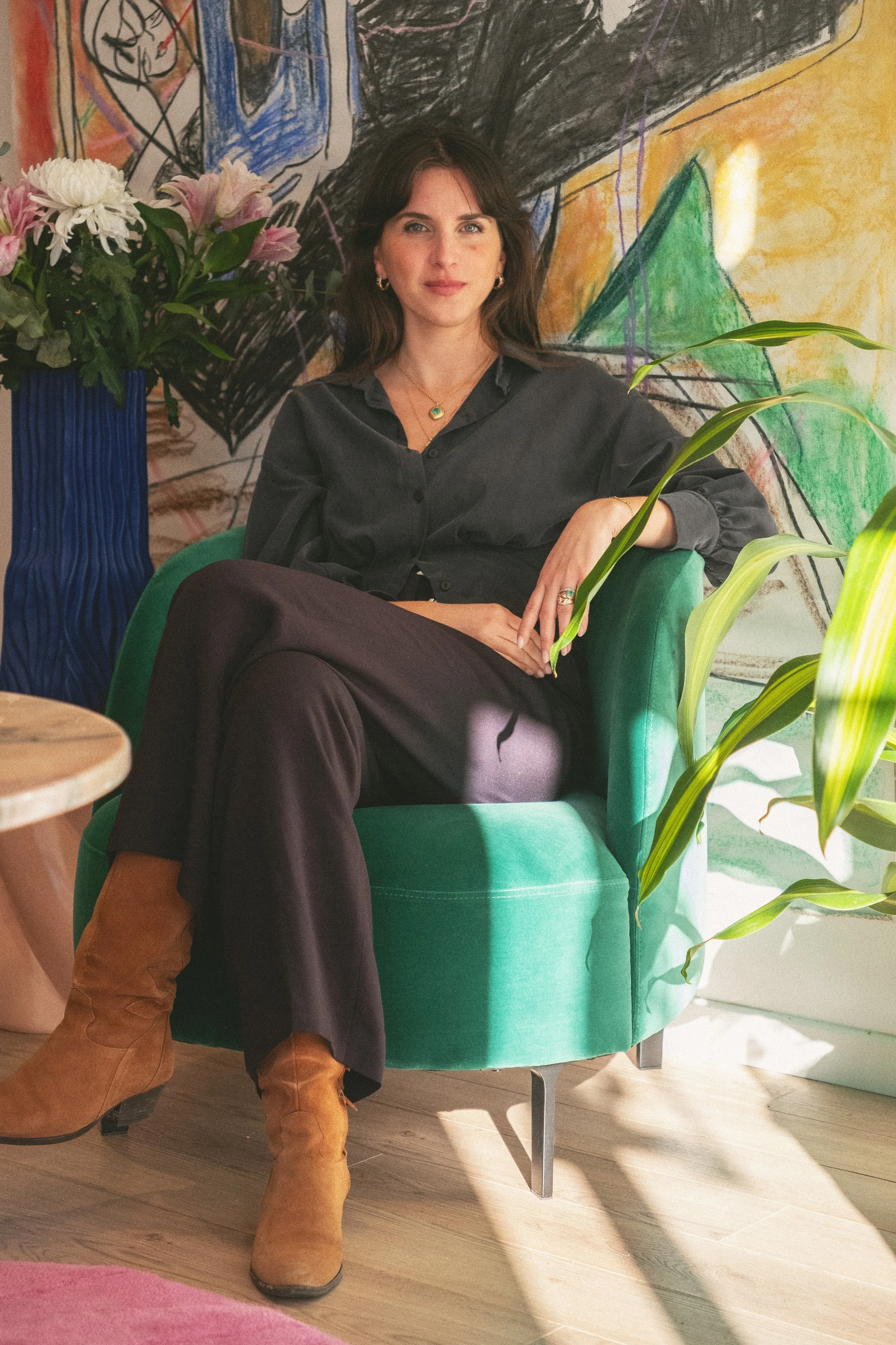 A young woman sitting in a green armchair, surrounded by plants, with an abstract painting in the background; she is wearing black clothes, a gold necklace, earrings, and beige suede boots.