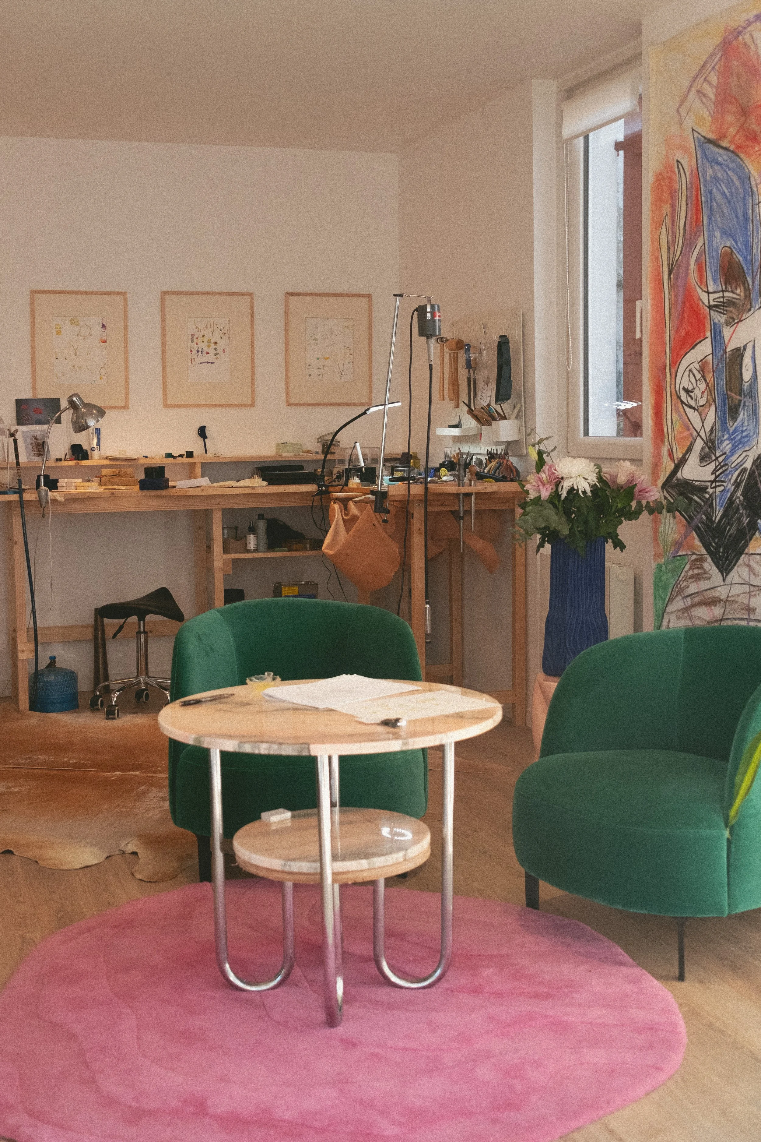 The interior of a room featuring a wooden table, two green chairs, a round pink rug, a large flower pot, a wall with artwork, and a workspace with various tools.