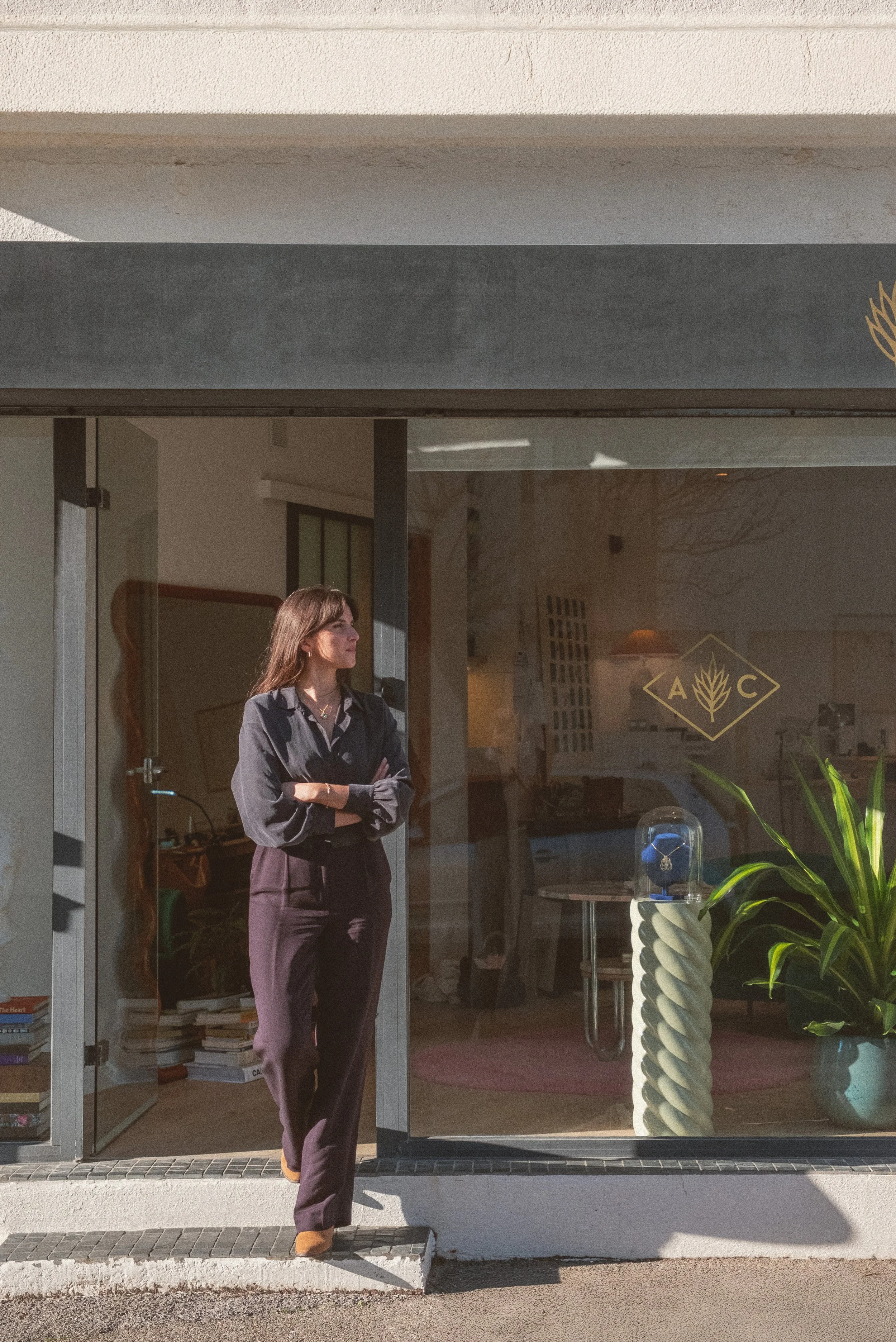 A young woman standing in front of a store window, wearing a dark gray blouse, purple pants, and brown shoes, with her arms crossed, in the bright light of late afternoon.