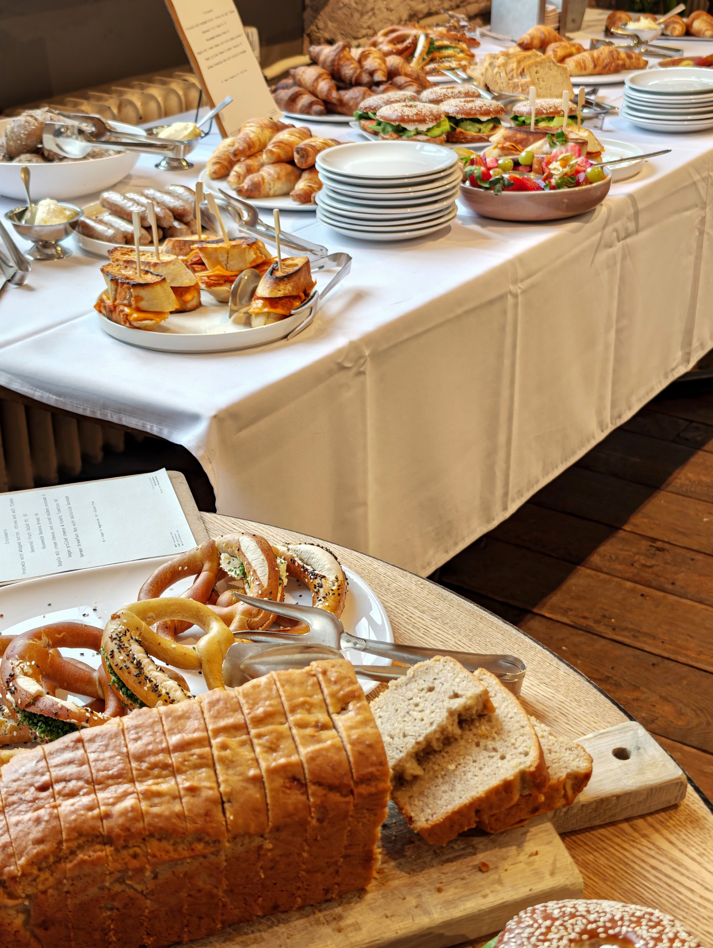 A table filled with sliced bread, pretzels, sandwiches, salad, croissants, and other baked goods at a buffet or breakfast setting.