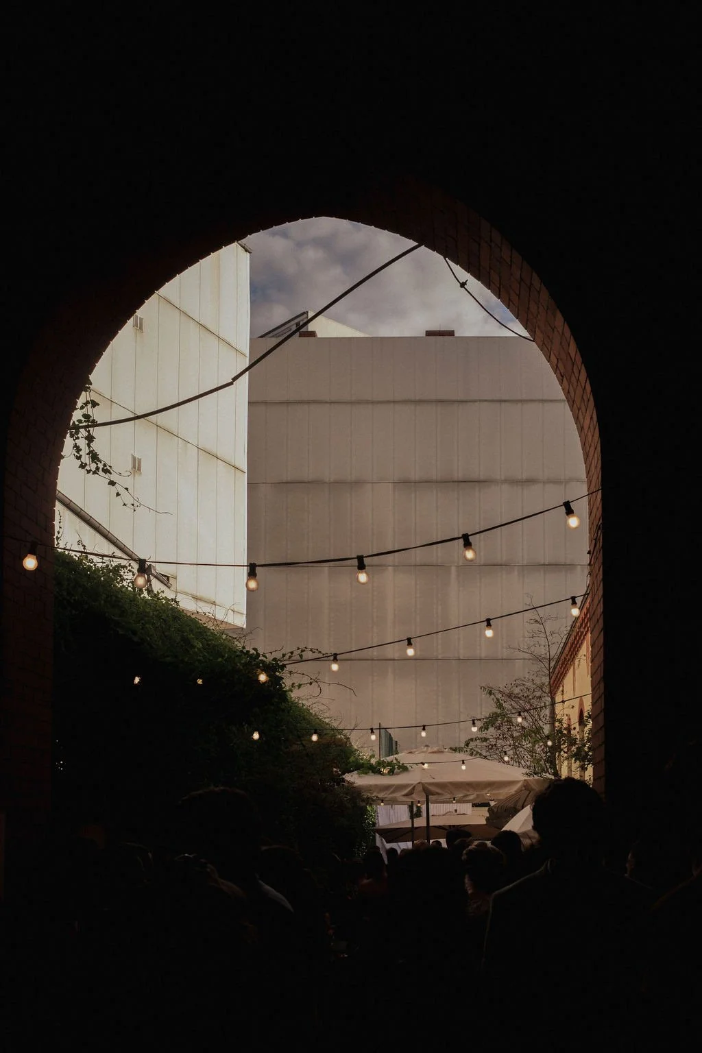 View through a dark archway showing outdoor patio with string lights, umbrellas, and a tall building in the background.