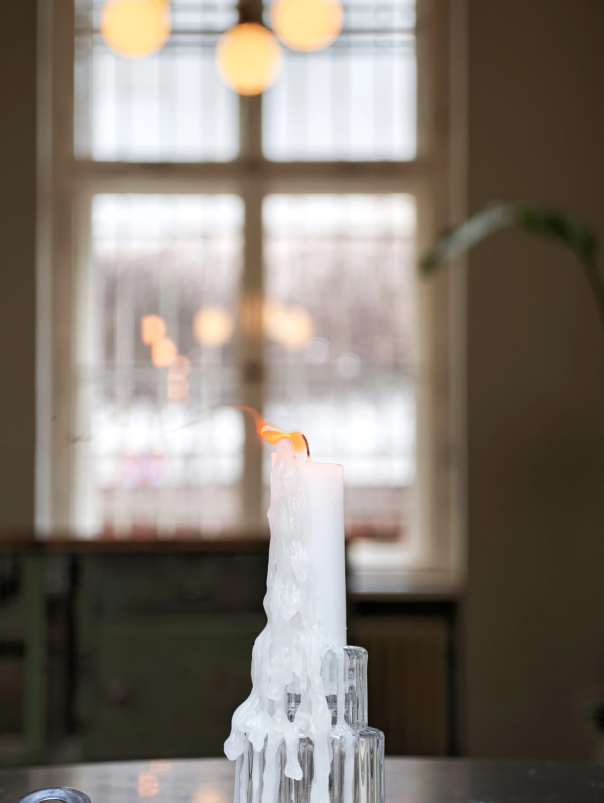 White candle melting on a glass holder in front of a window with blurred hanging lights in the background.