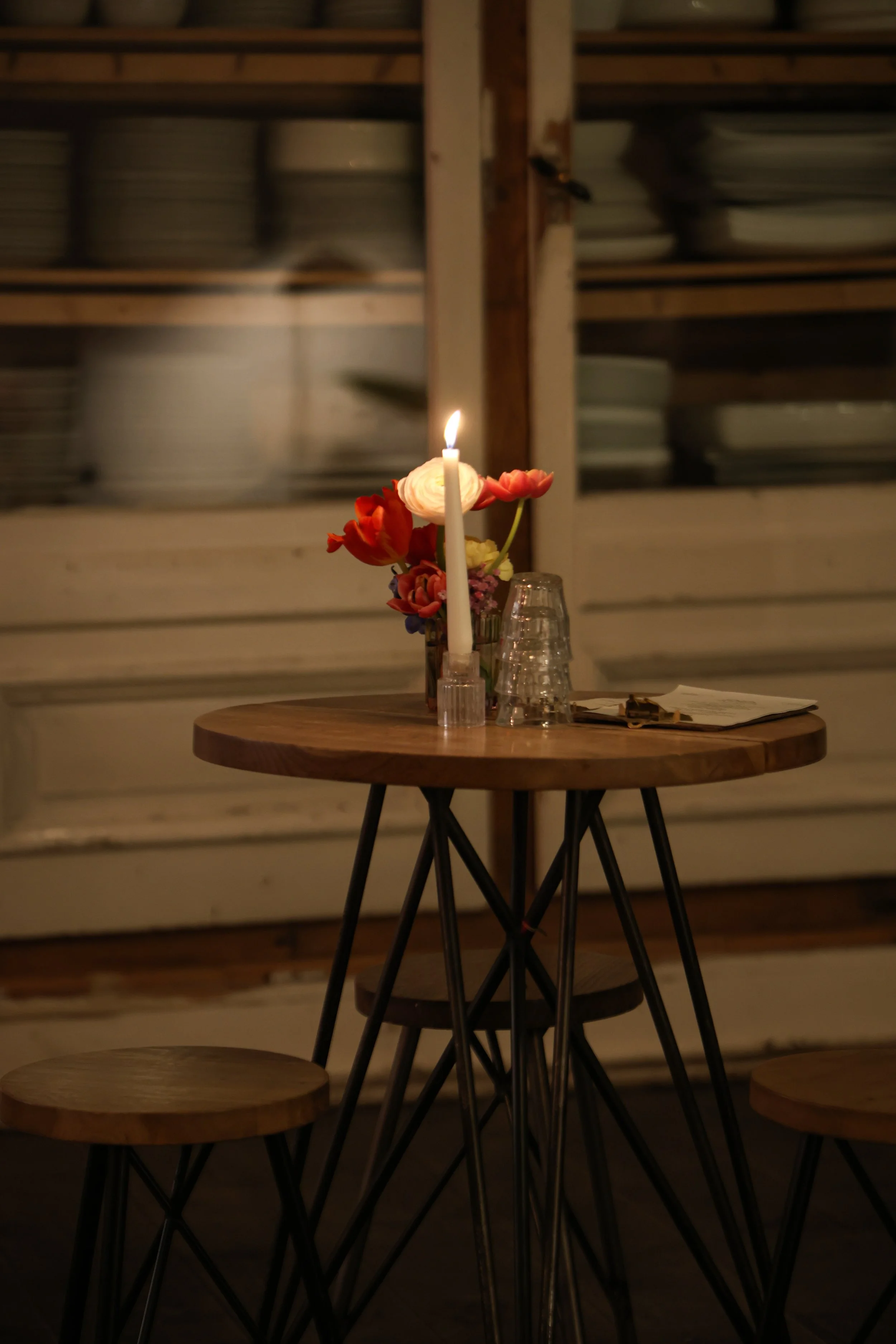 Wooden table with a flower bouquet, a lit candle, and upside-down glasses. In the background, shelves with plates and bowls.
