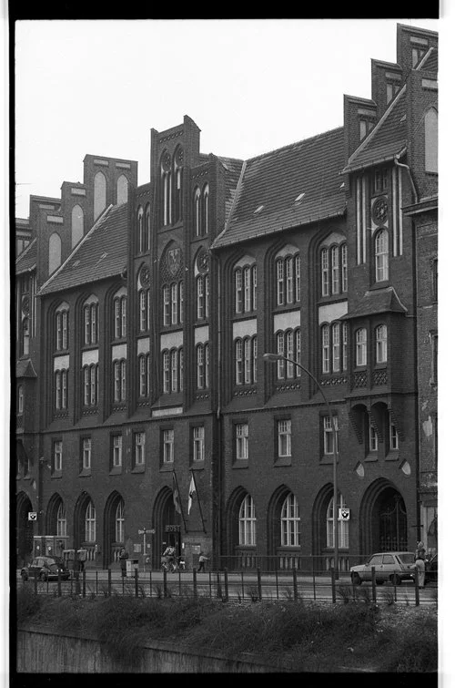 Black and white photo of a multi-story brick building with Gothic architectural features, including pointed arches, tall narrow windows, and decorative elements on the upper parts, located along a street with cars and pedestrians.