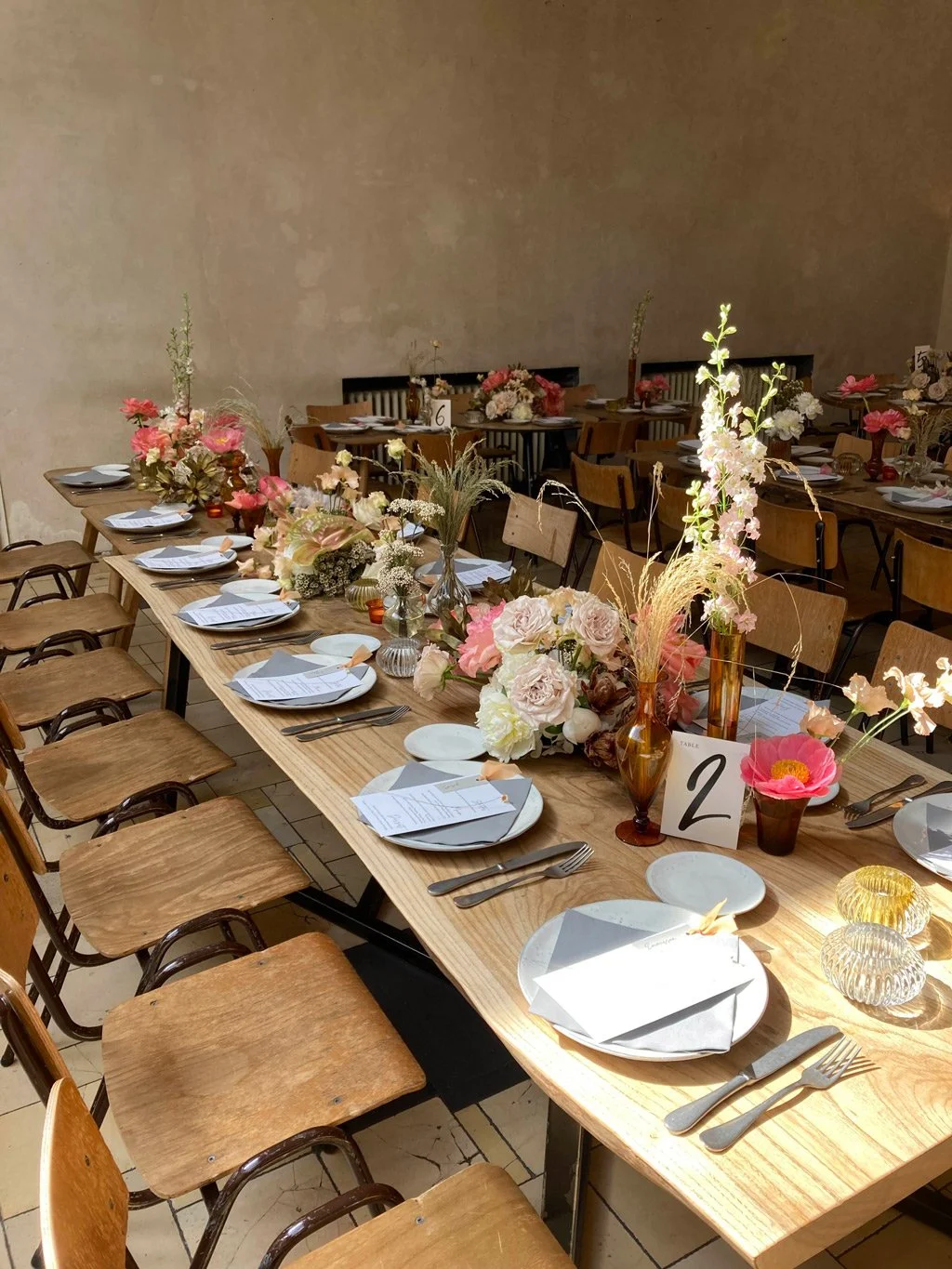 A long wooden table set for an event with floral arrangements, plates, silverware, and napkins, with chairs on both sides and additional tables in the background with similar decor.