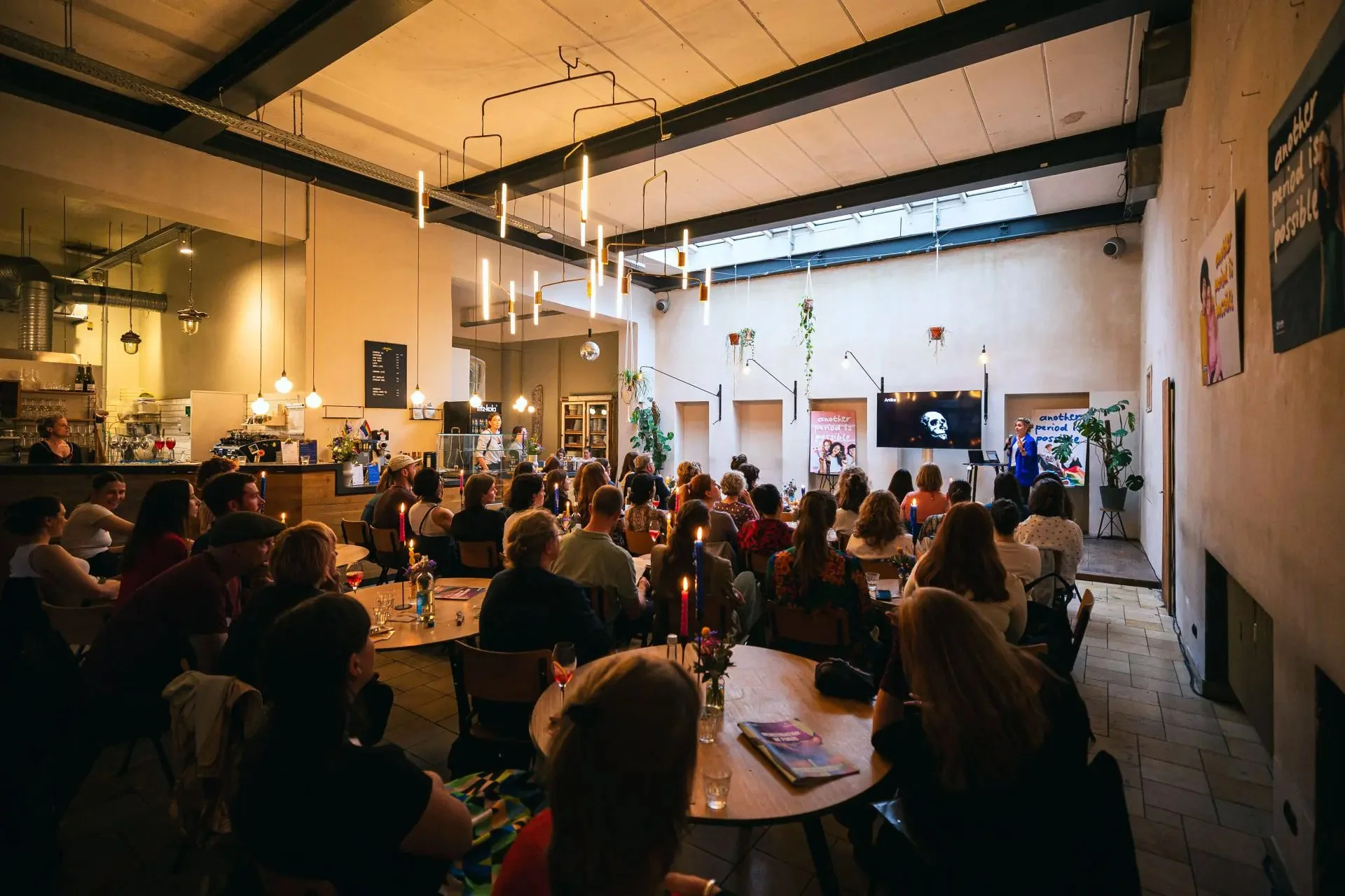 A crowded indoor venue with people attending a live event. There are tables with candles and flowers, a bar area on the left, and a stage with a presenter on the right. The room has high ceilings with modern hanging lights and a skylight.