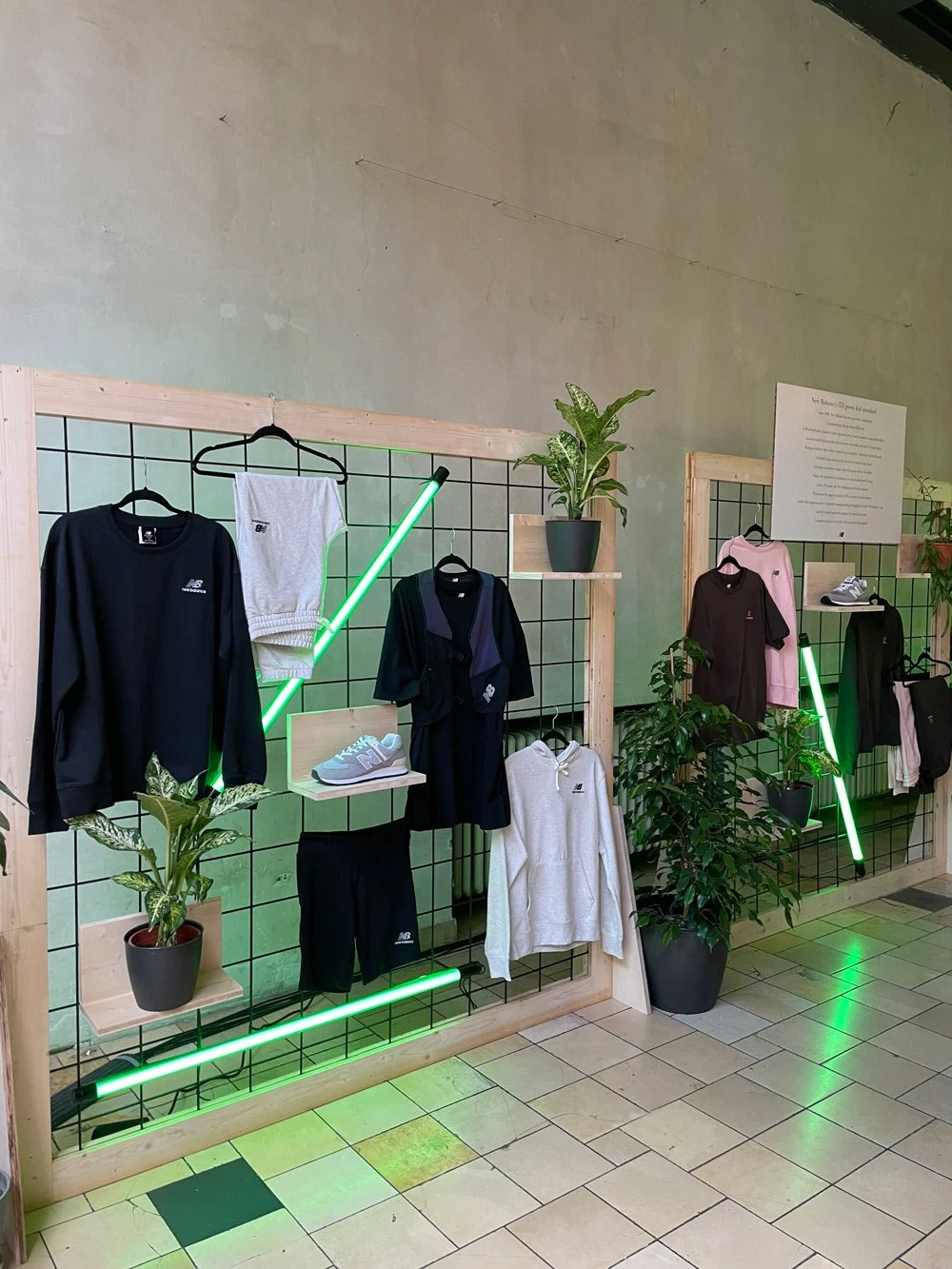 Display of athletic clothing and sneakers on a wire grid with wooden framing, green neon lights, and potted plants inside a store.