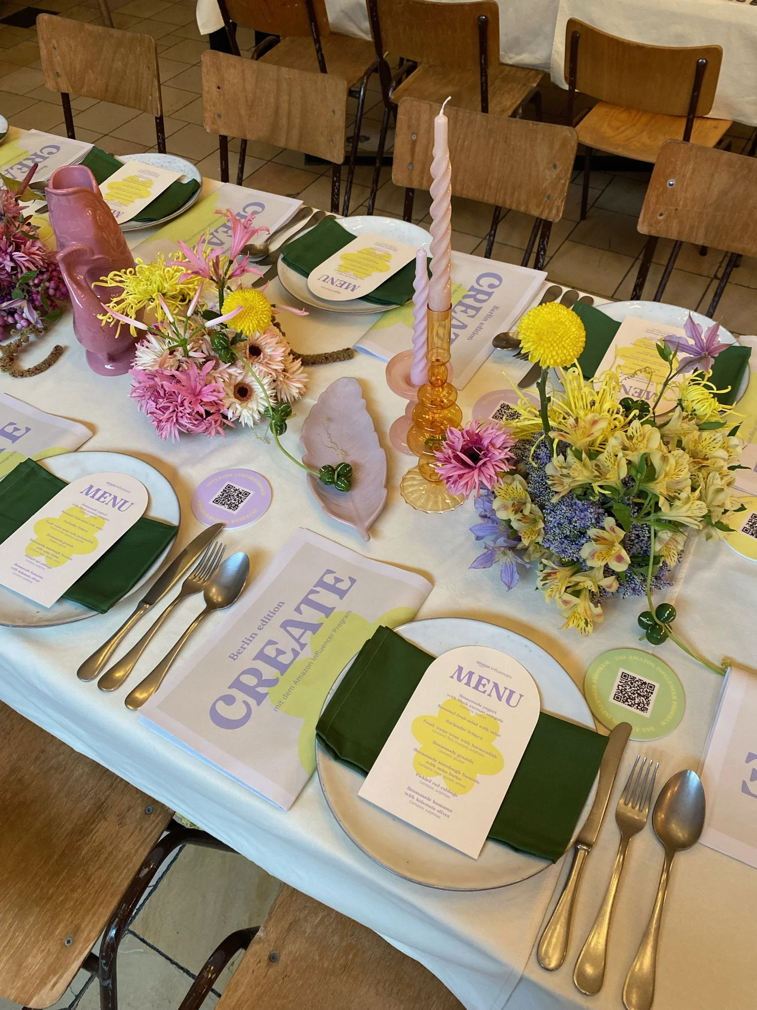 A dining table set with plates, silverware, and menus for a restaurant or event. The table has floral centerpieces, candles, and napkins. Wooden chairs are arranged around the table.