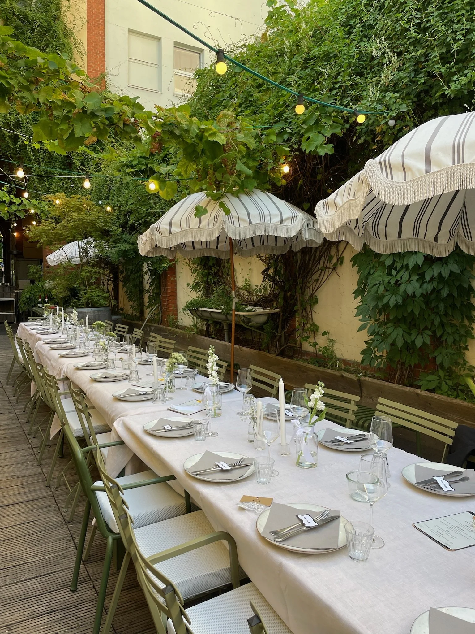 Long outdoor dining table set with white tablecloth, plates, glasses, and silverware, surrounded by beige chairs, under striped umbrellas, in a lush green garden with string lights.