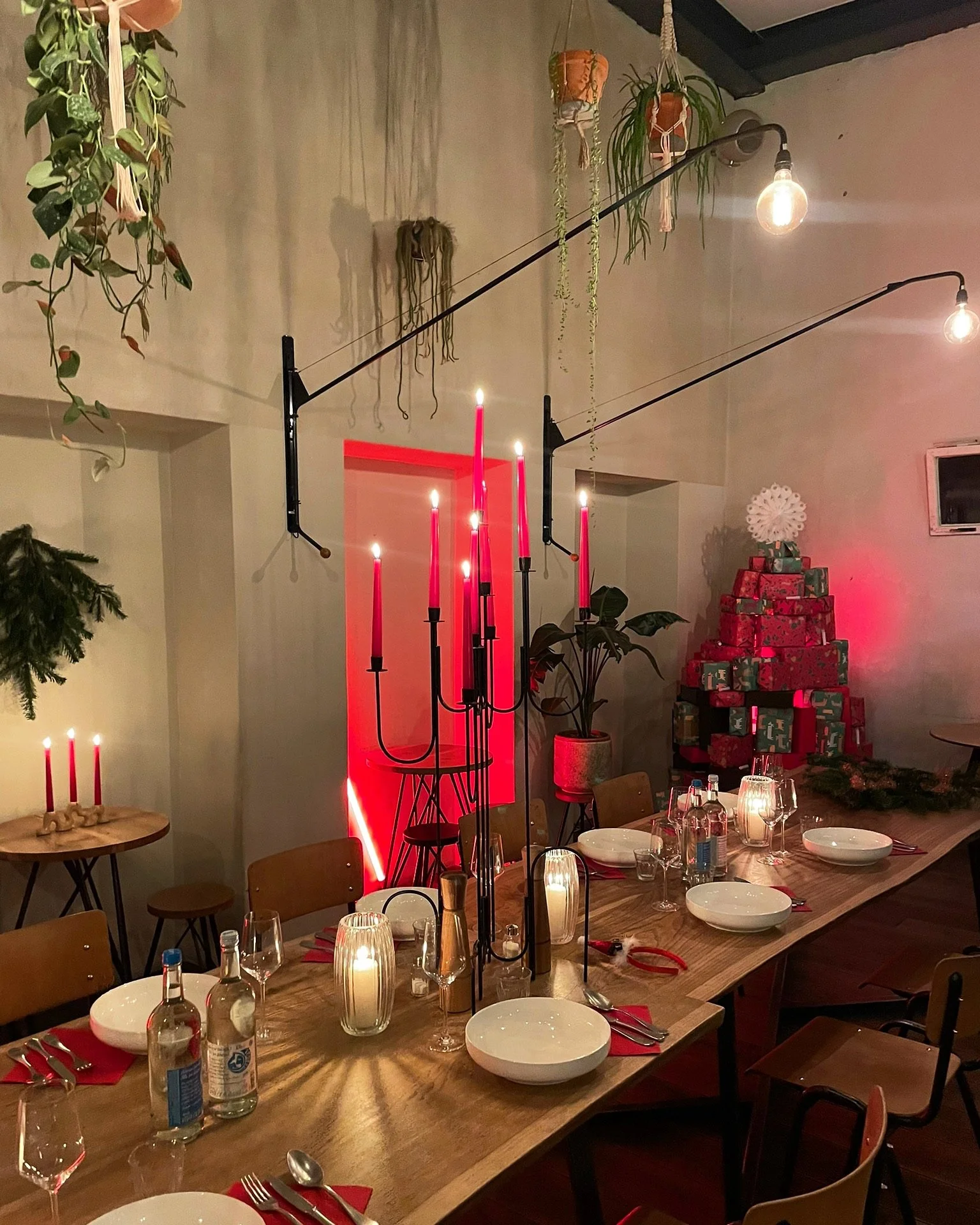 A dining table decorated with lit candles, wine glasses, plates, and cutlery, with a backdrop of hanging plants, a stack of wrapped Christmas presents, and warm ambient lighting.