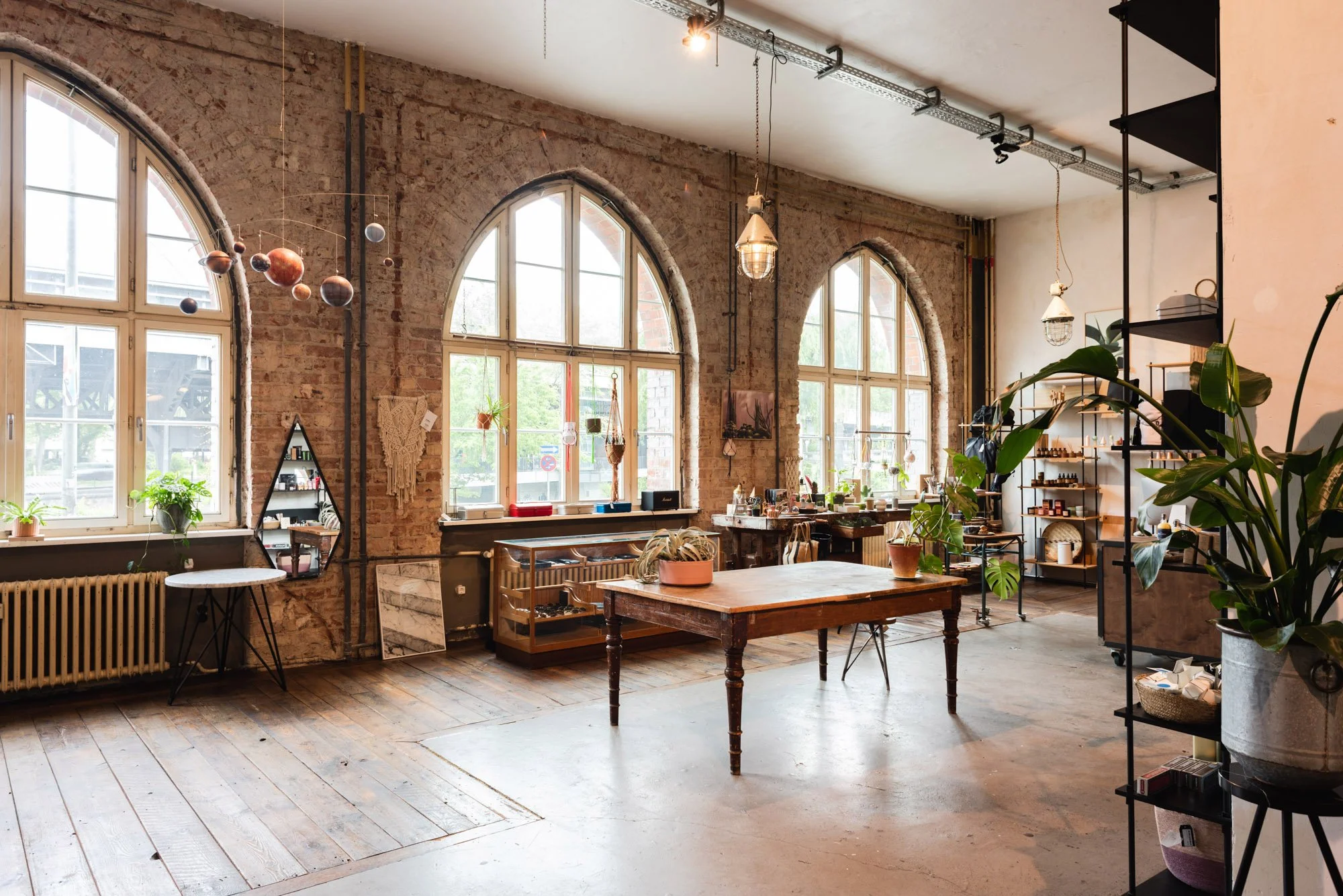 Interior of a cozy brick-walled space with large arched windows, wooden floors, and a variety of plants and decorative items on tables and shelves, illuminated by hanging and ceiling lights.