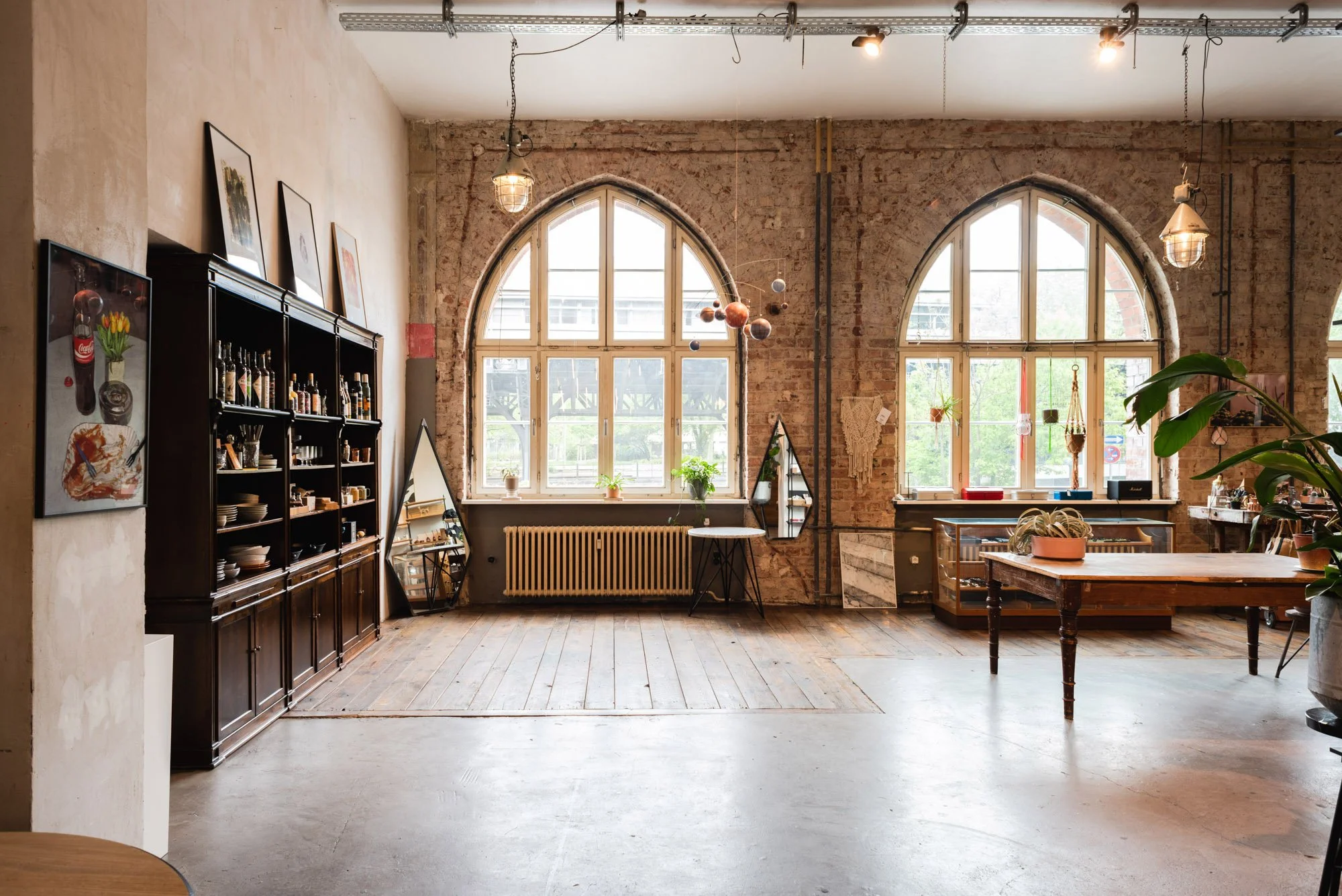 Interior of a spacious room with large arched windows, exposed brick walls, hanging lights, and wooden furniture, decorated with plants and artwork.