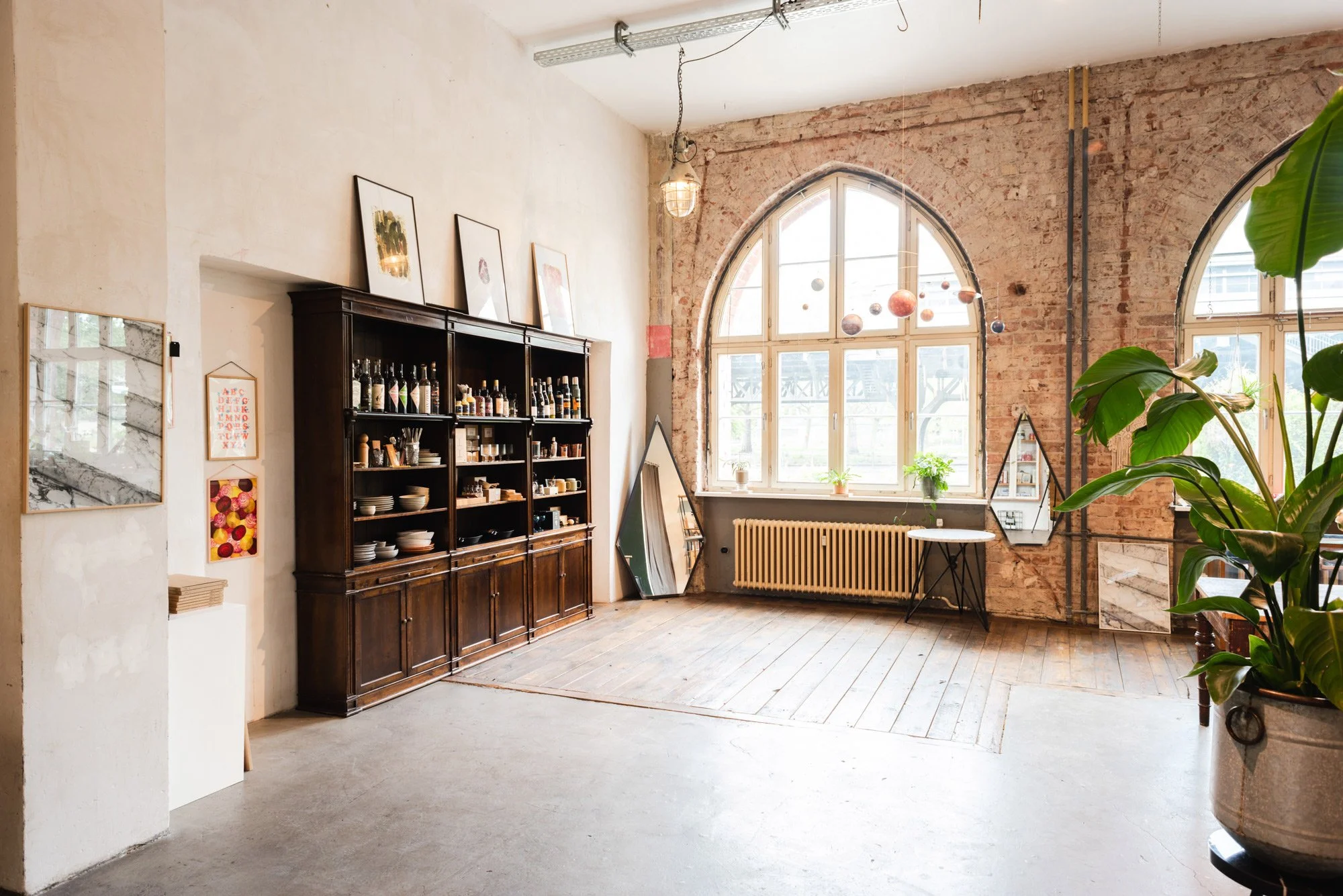 Interior of a spacious room with exposed brick walls, large arched windows allowing natural light, a dark wooden shelving unit filled with dishes and bottles, framed artwork, and potted plants.