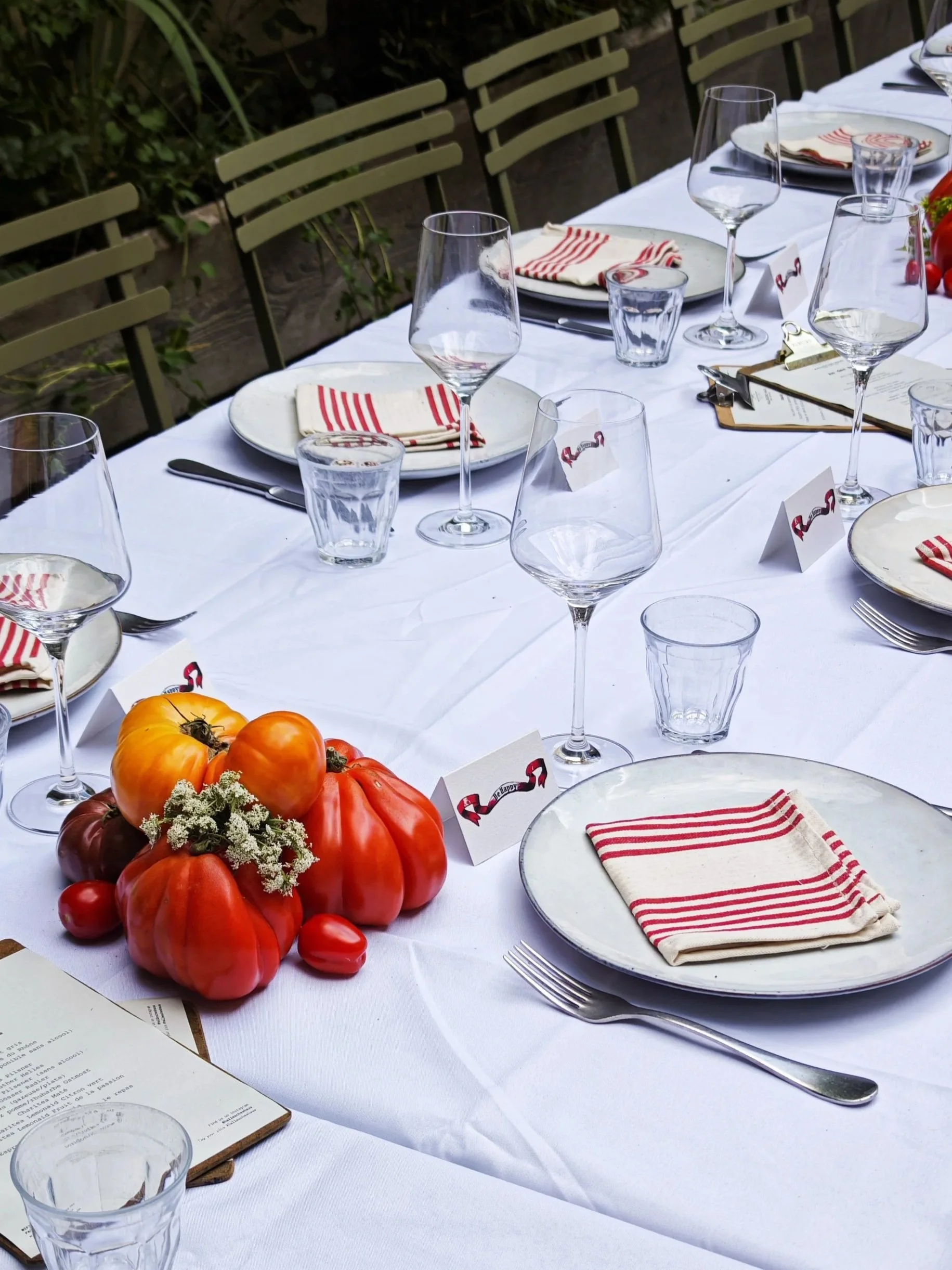 A long dining table set outdoors with white tablecloth, empty plates with red and white striped napkins, wine glasses, water glasses, and silverware. A centerpiece of heirloom tomatoes and cherry tomatoes on a plate. Place cards and menus are on the 