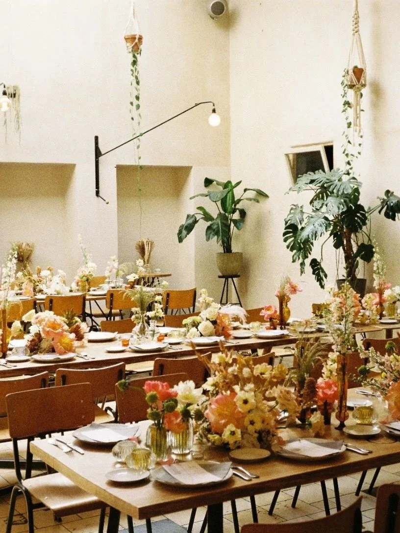 Long wooden tables set for a celebration with floral centerpieces, surrounded by wooden chairs, in a bright room with plants and hanging planters.