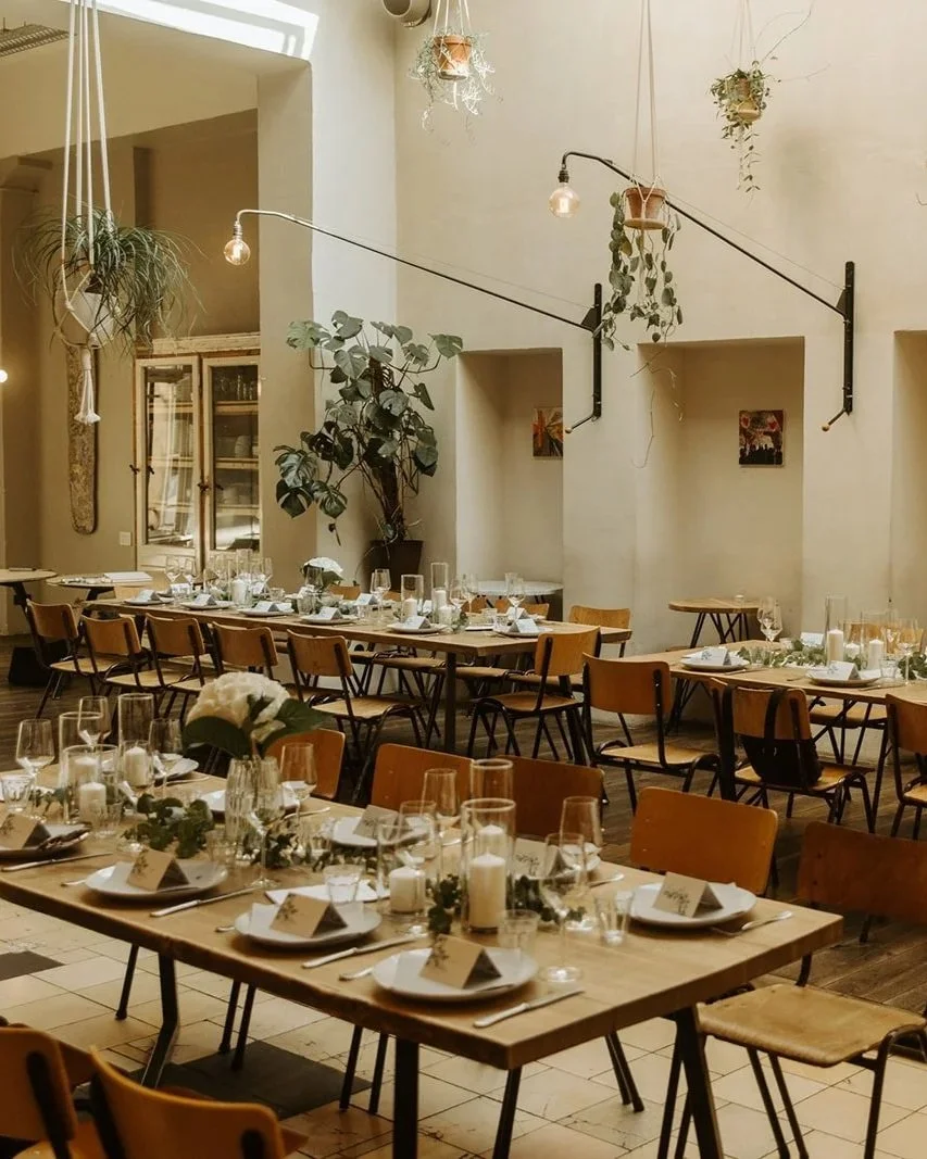 A dining area set up for a special event with long wooden tables decorated with candles, flowers, and place settings, surrounded by wooden chairs, and adorned with hanging plants and minimalistic wall decorations.
