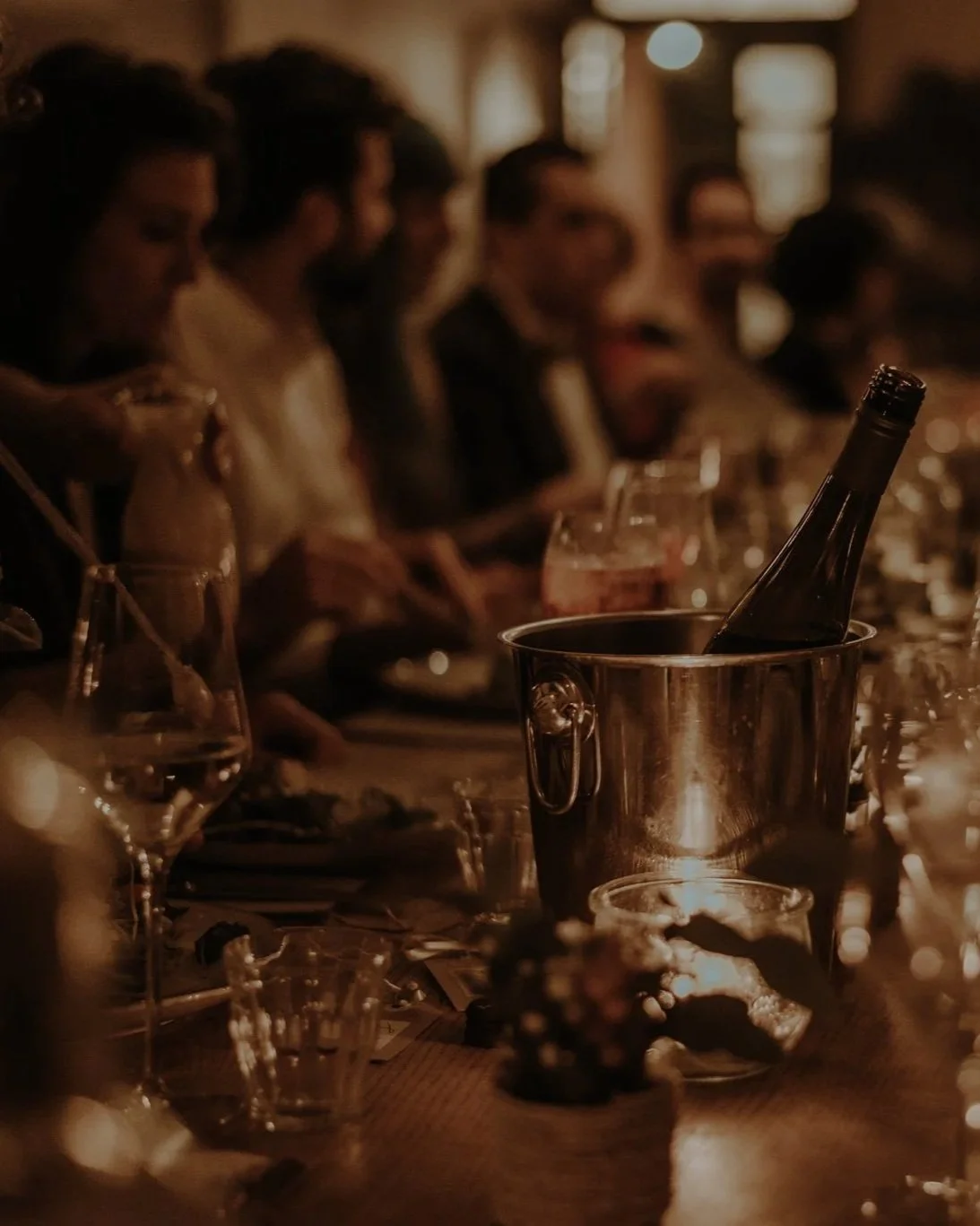 A dimly lit dinner table with people socializing, drinks, and a wine bottle in an ice bucket.