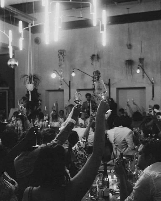 People sitting at tables during a celebration, raising glasses in a toast, with a band playing on stage in the background, illuminated by hanging lights.