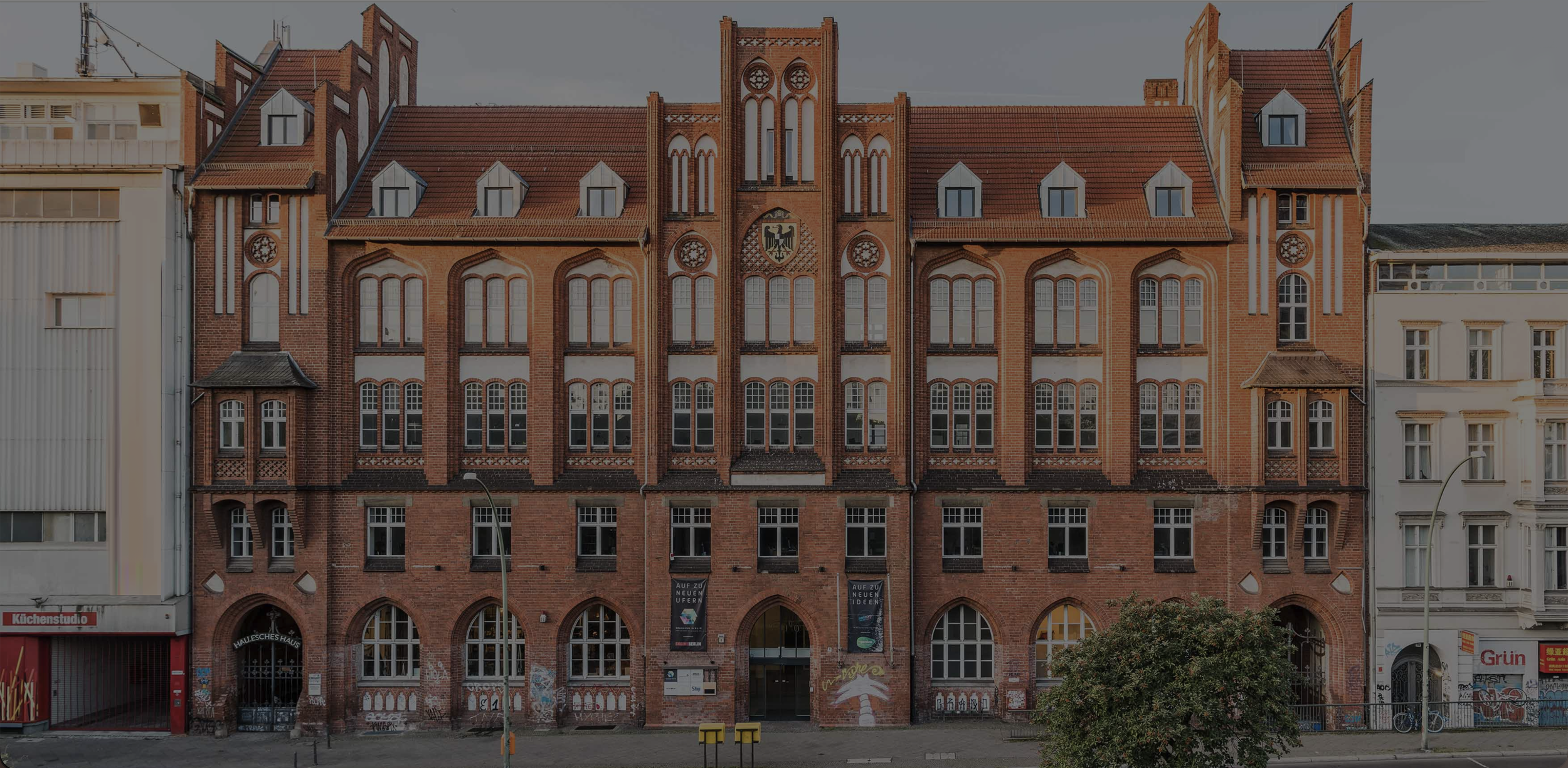 Historic red brick building with Gothic architectural details, large arched windows, and a central coat of arms emblem, located between modern structures.