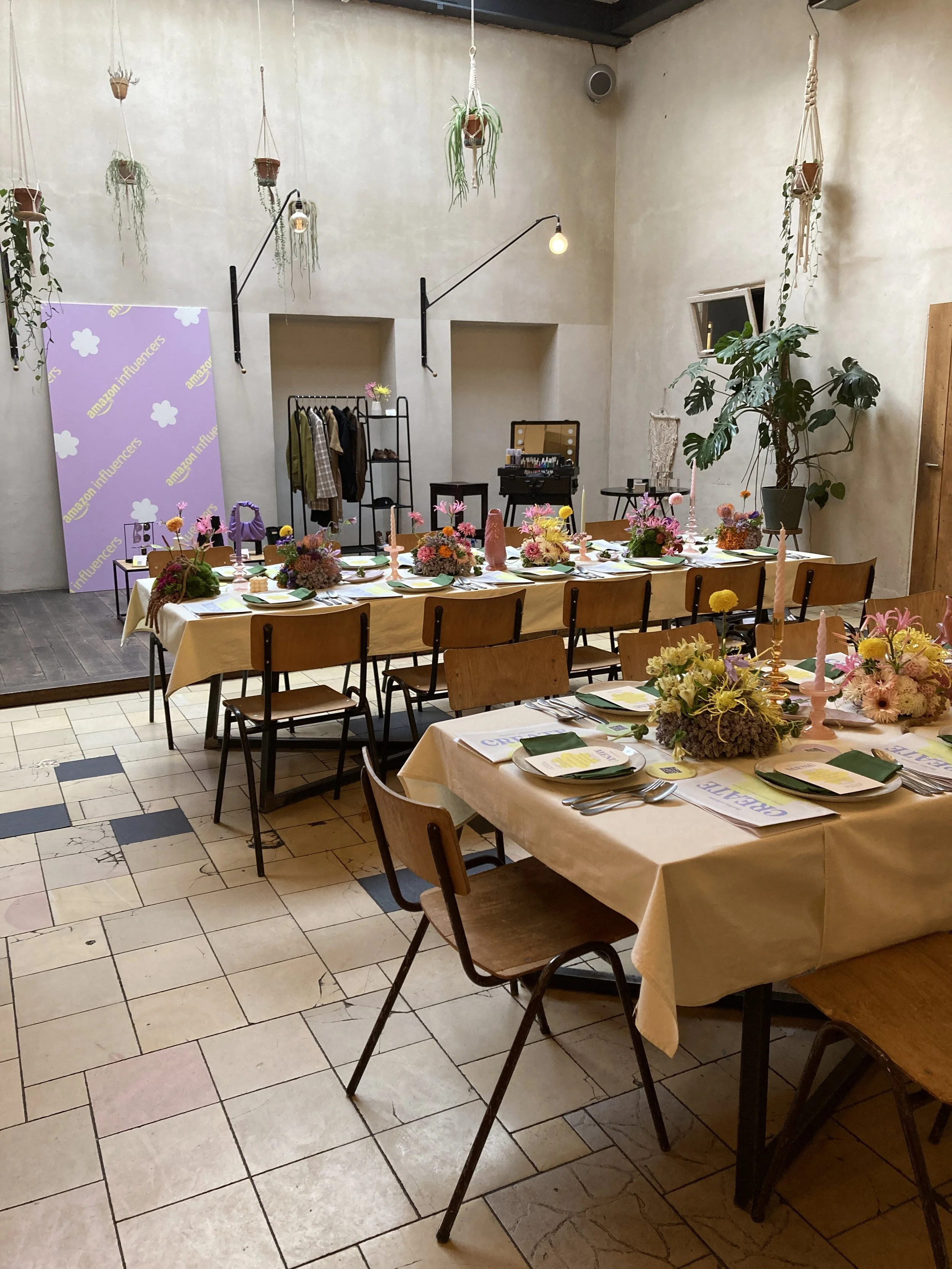 A dining area decorated for an event with long tables covered in beige tablecloths, floral centerpieces, pink candles, and place settings. The room has hanging plants from the ceiling, a large green plant near a small window, and a coat rack with clo