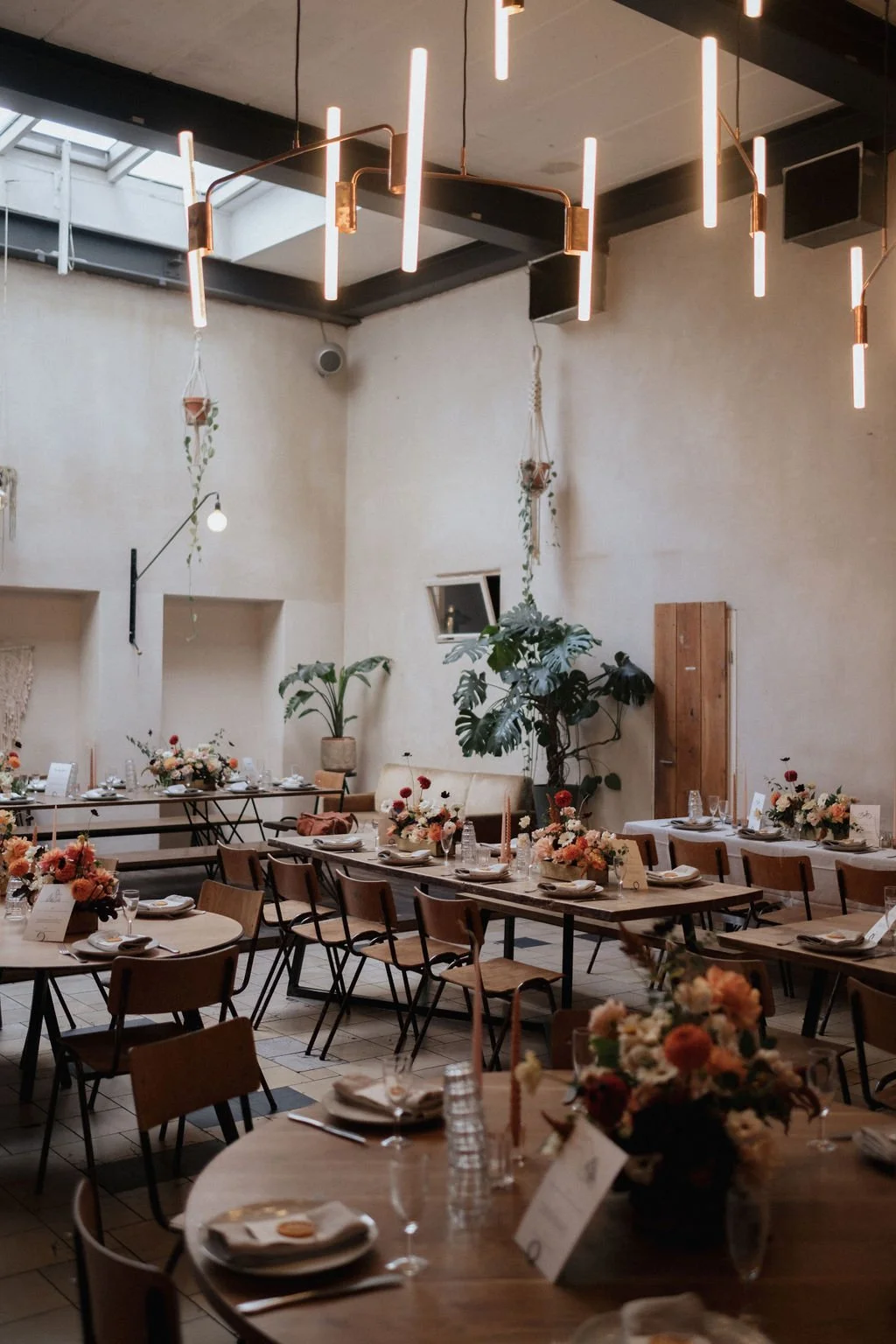 Interior of a decorated event space with tables set for a meal, floral centerpieces, and modern hanging light fixtures.