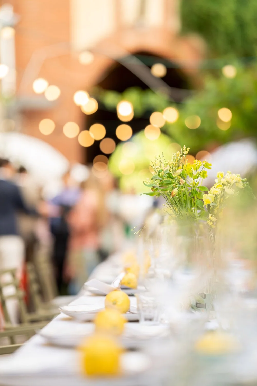 Decorative outdoor dining setup with flowers, lemons, and blurred string lights and people in the background.