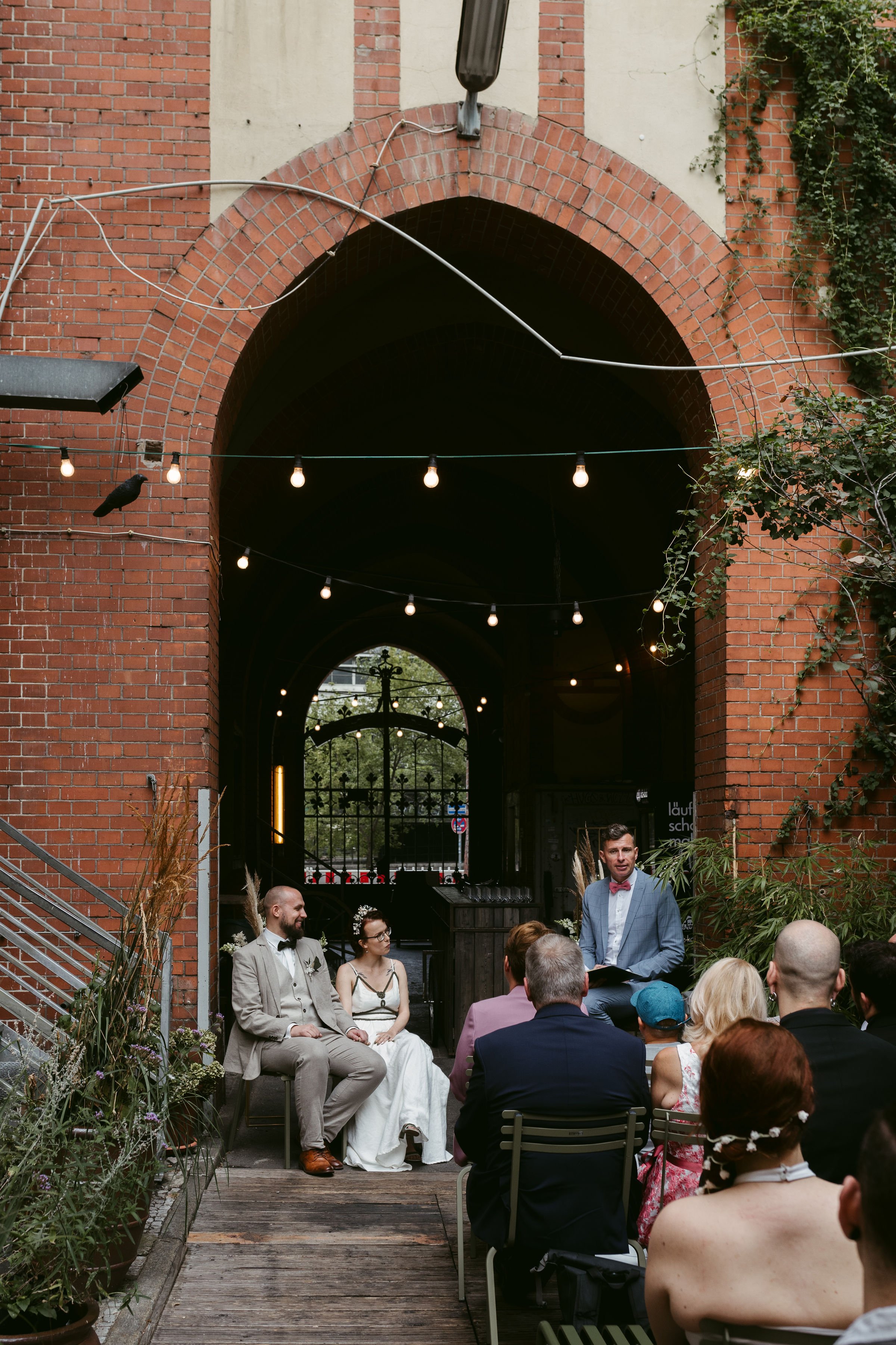A wedding ceremony taking place under a brick arch with string lights, with a group of guests seated and a man in a suit speaking at the event.