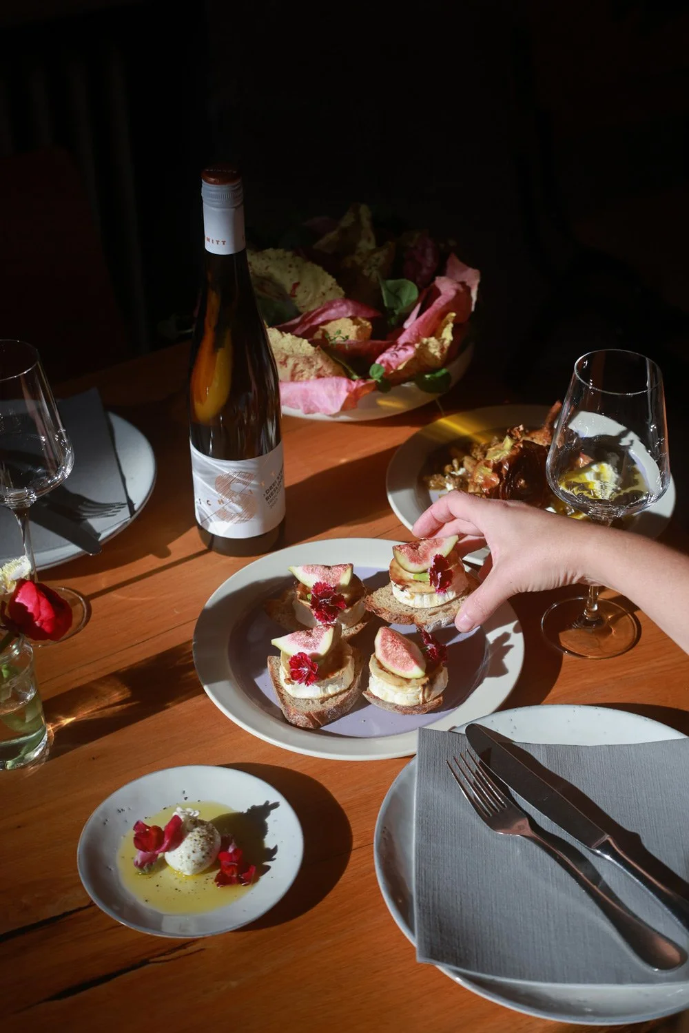 A person serving three small open-faced sandwiches topped with fig slices, banana slices, and edible flowers, on a white plate. Other food items include a bowl of roasted meat, a wine glass with white wine, a water glass, a bottle of beer, a small dish with a dollop of cheese or cream garnished with edible flowers, and a bowl of bread or chips wrapped in pink paper. The table is set with silverware and napkins, and the atmosphere appears to be a dinner setting.