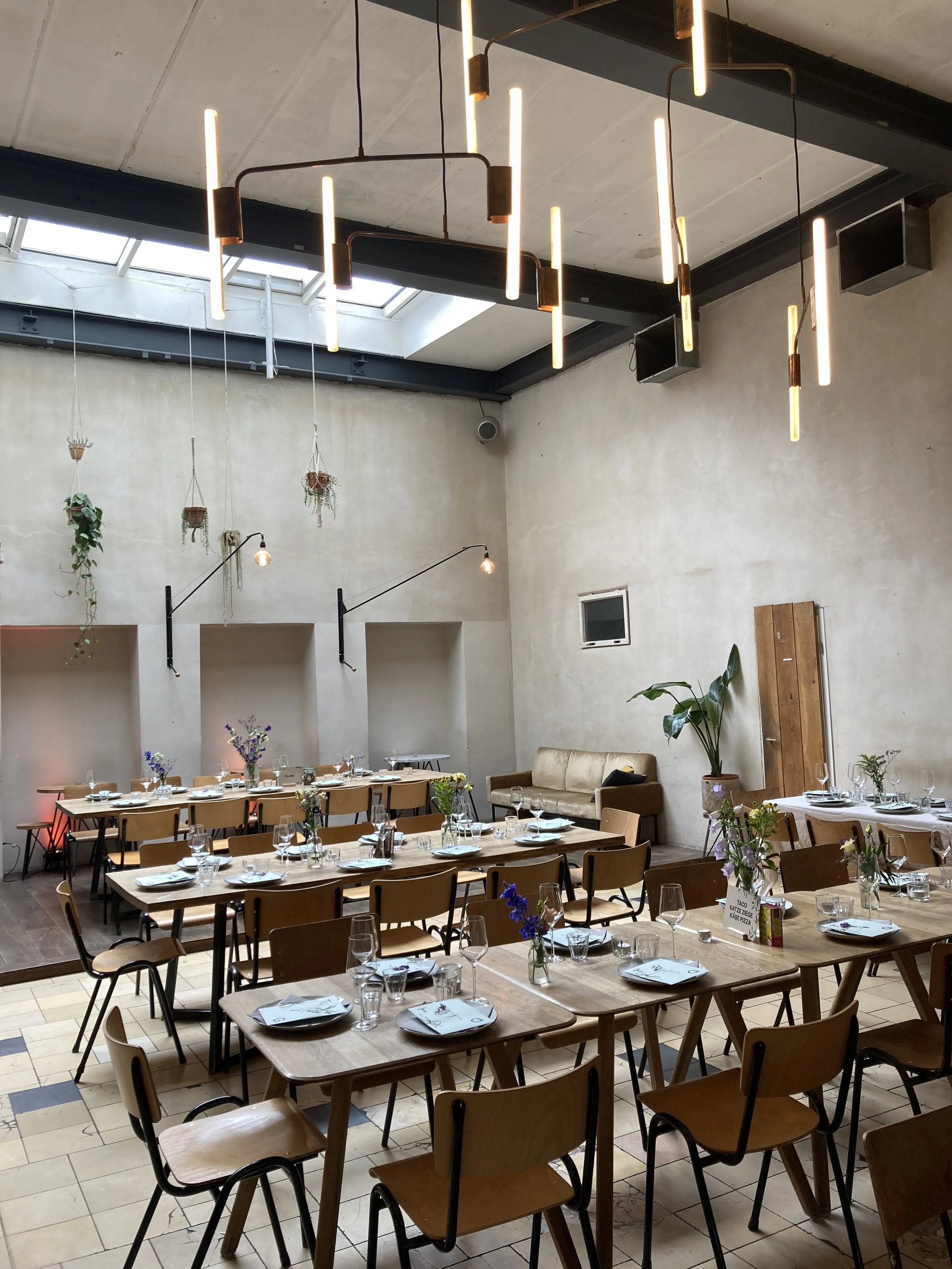 Interior of a modern restaurant or cafe with wooden tables, chairs, and set diningware. Hanging lights and potted plants adorn the space, and there is a skylight overhead.