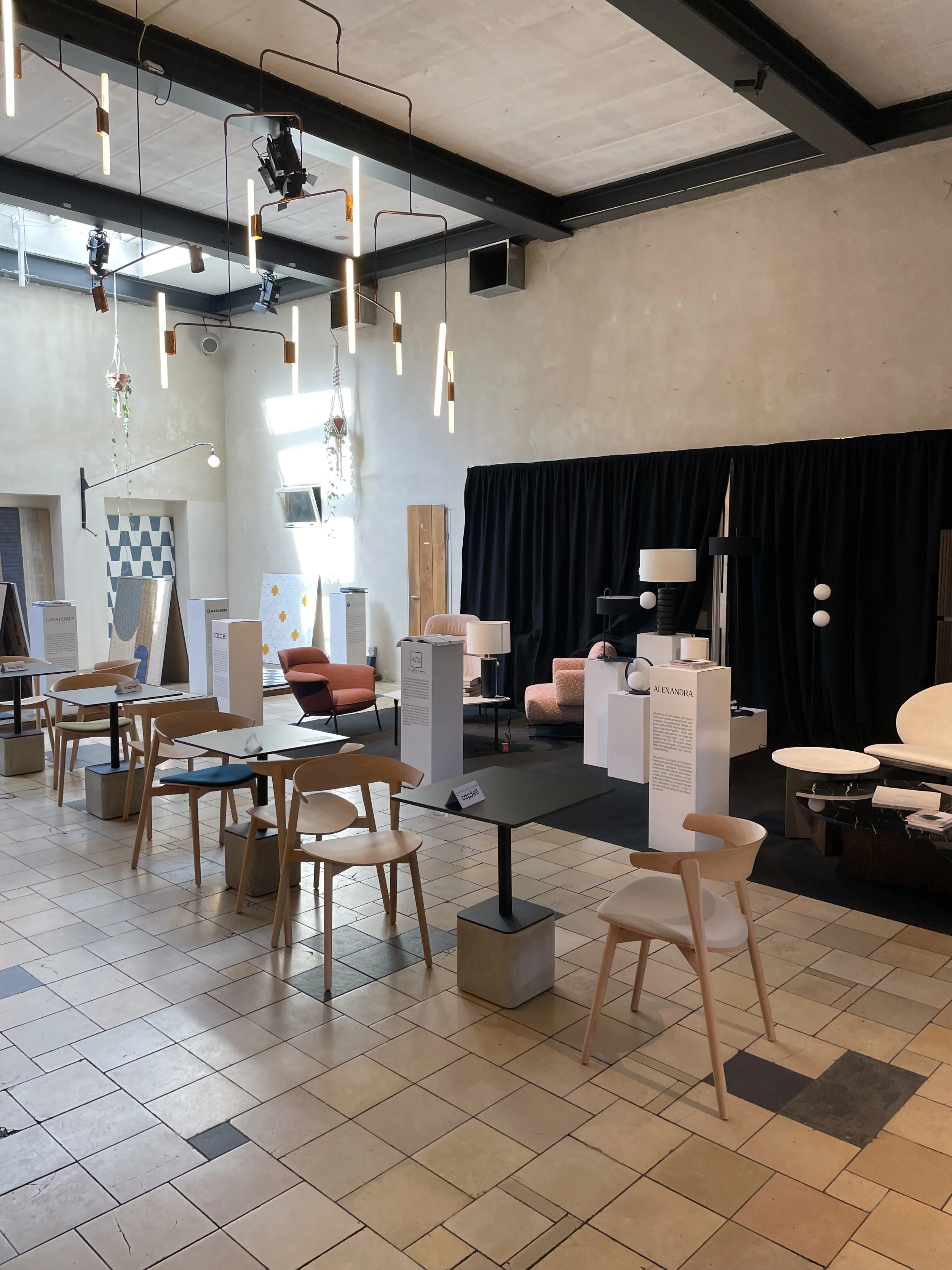 A modern furniture showroom with various chairs, tables, and lamps on display, featuring a high ceiling with contemporary hanging lights.