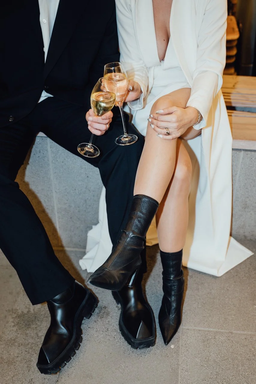Two people, one in a black suit and the other in a white dress and black boots, sitting close together and holding glasses of champagne.