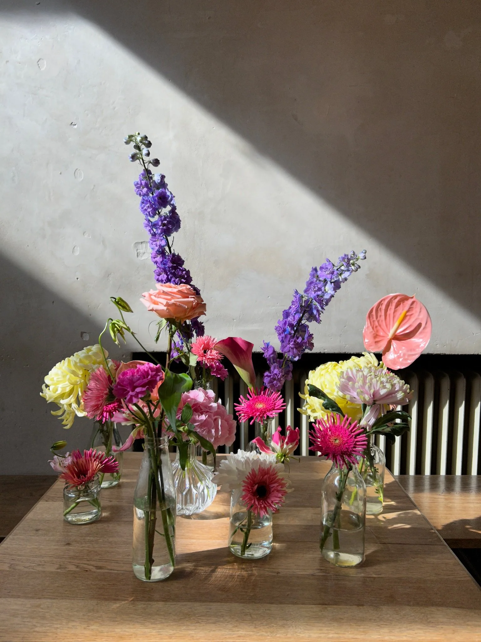 A collection of colorful flowers in vases on a wooden table, with sunlight casting shadows on a plain wall behind them.