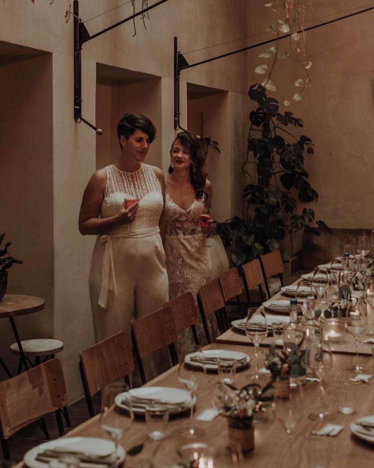 Two women in elegant dresses standing beside a long, decorated dining table in a warmly lit room, with plates, glasses, and floral centerpieces.