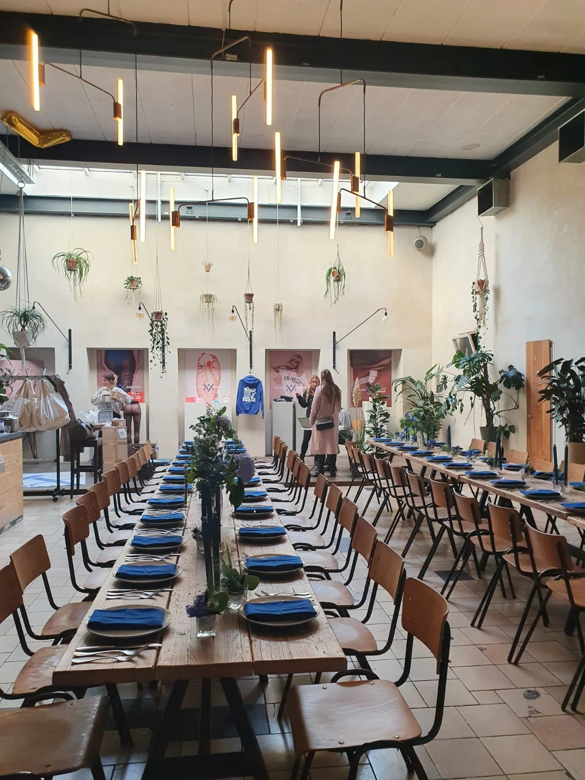 Interior of a restaurant or event space with long wooden tables set for a meal, decorated with flowers and surrounded by wooden chairs, with people preparing and arranging the space.
