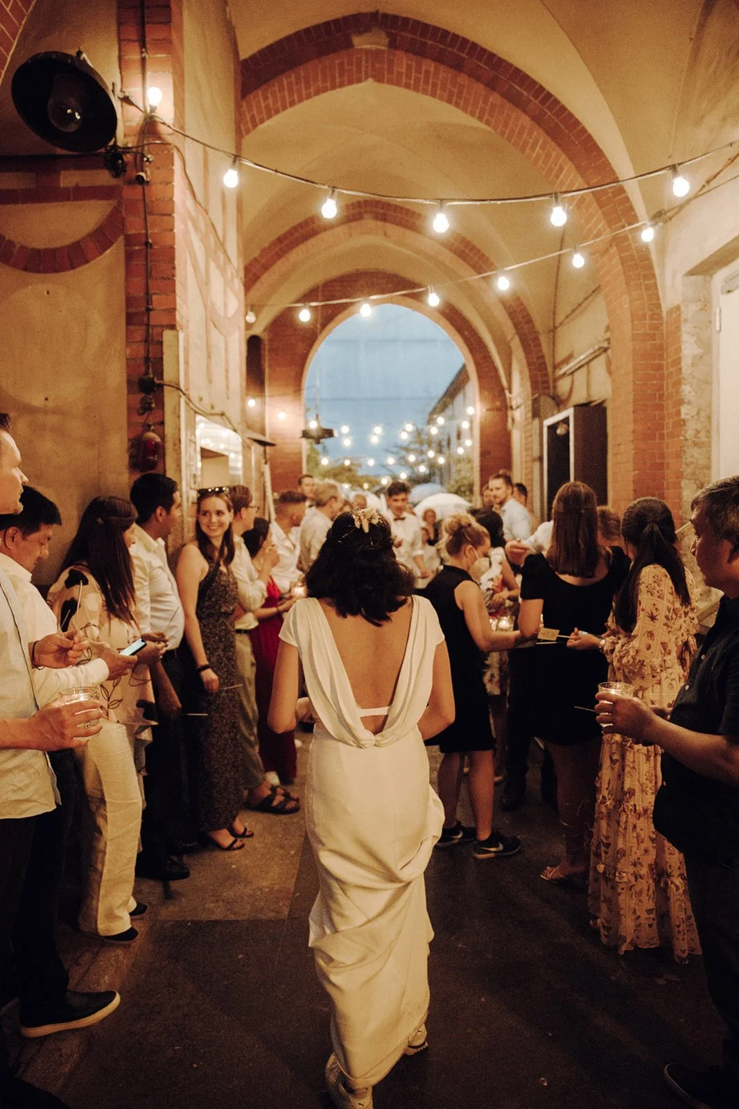 A woman in a white dress walking through a crowd of people at an outdoor evening event under string lights in an arched brick alleyway.