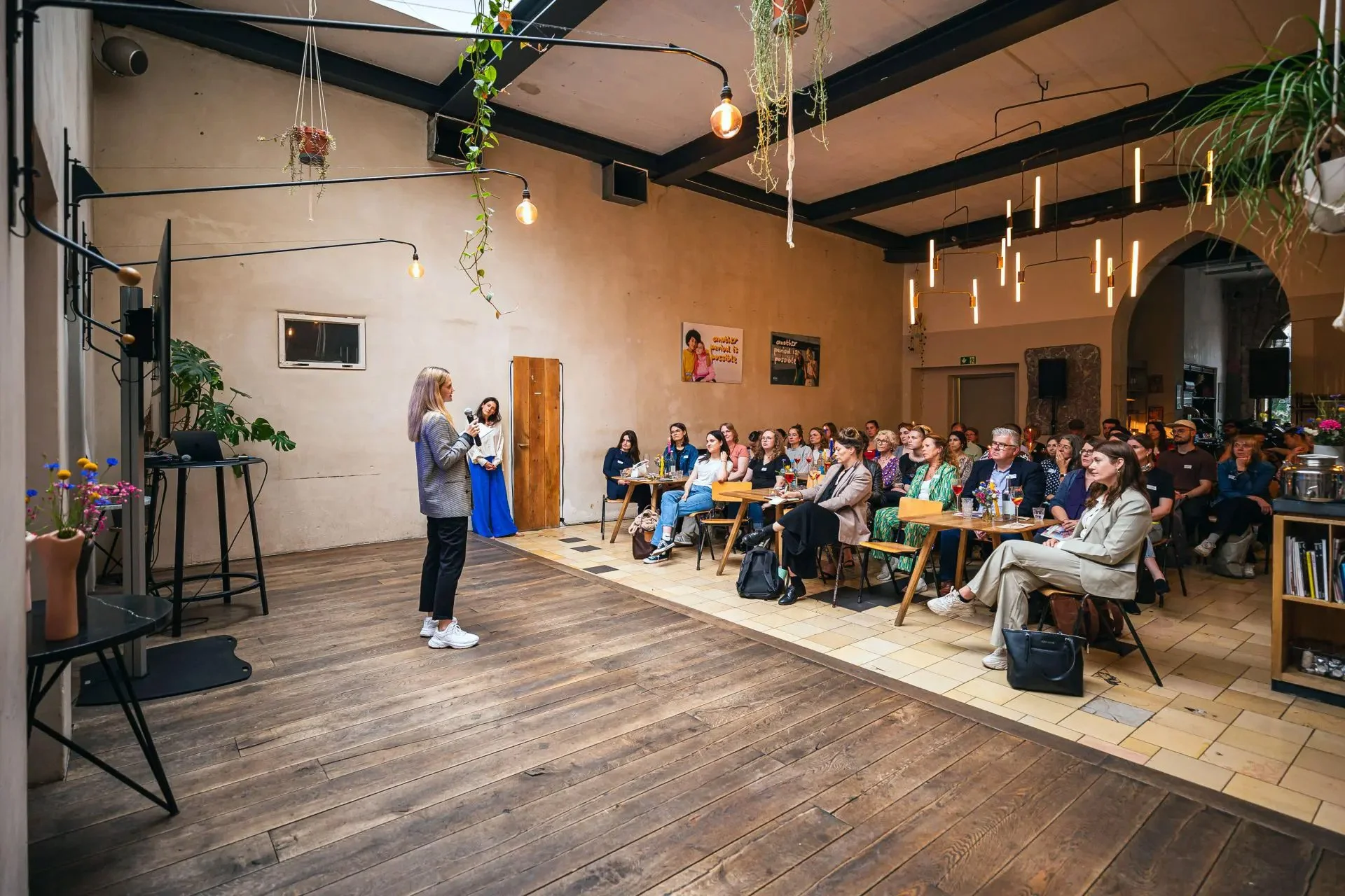 A woman in a gray blazer and white sneakers giving a presentation to a seated audience in a large, well-lit room with high ceilings, hanging lights, and decorative plants.